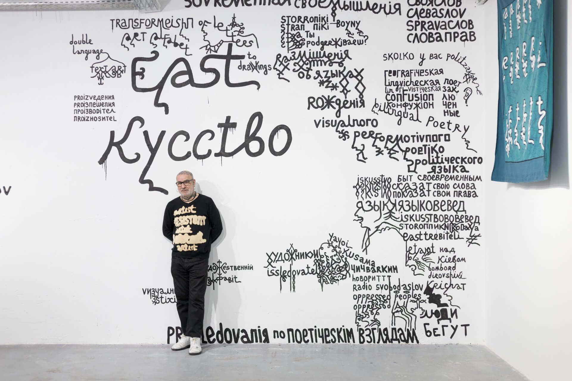 Une personne portant des lunettes se tient devant un mur blanc couvert de grands mots et phrases manuscrits en noir, en russe et en anglais, avec un textile bleu suspendu à droite.