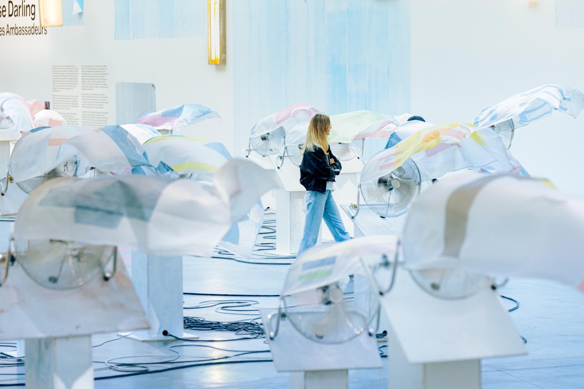 A woman stands among an art installation featuring fabric draped over fans in a brightly lit gallery space.