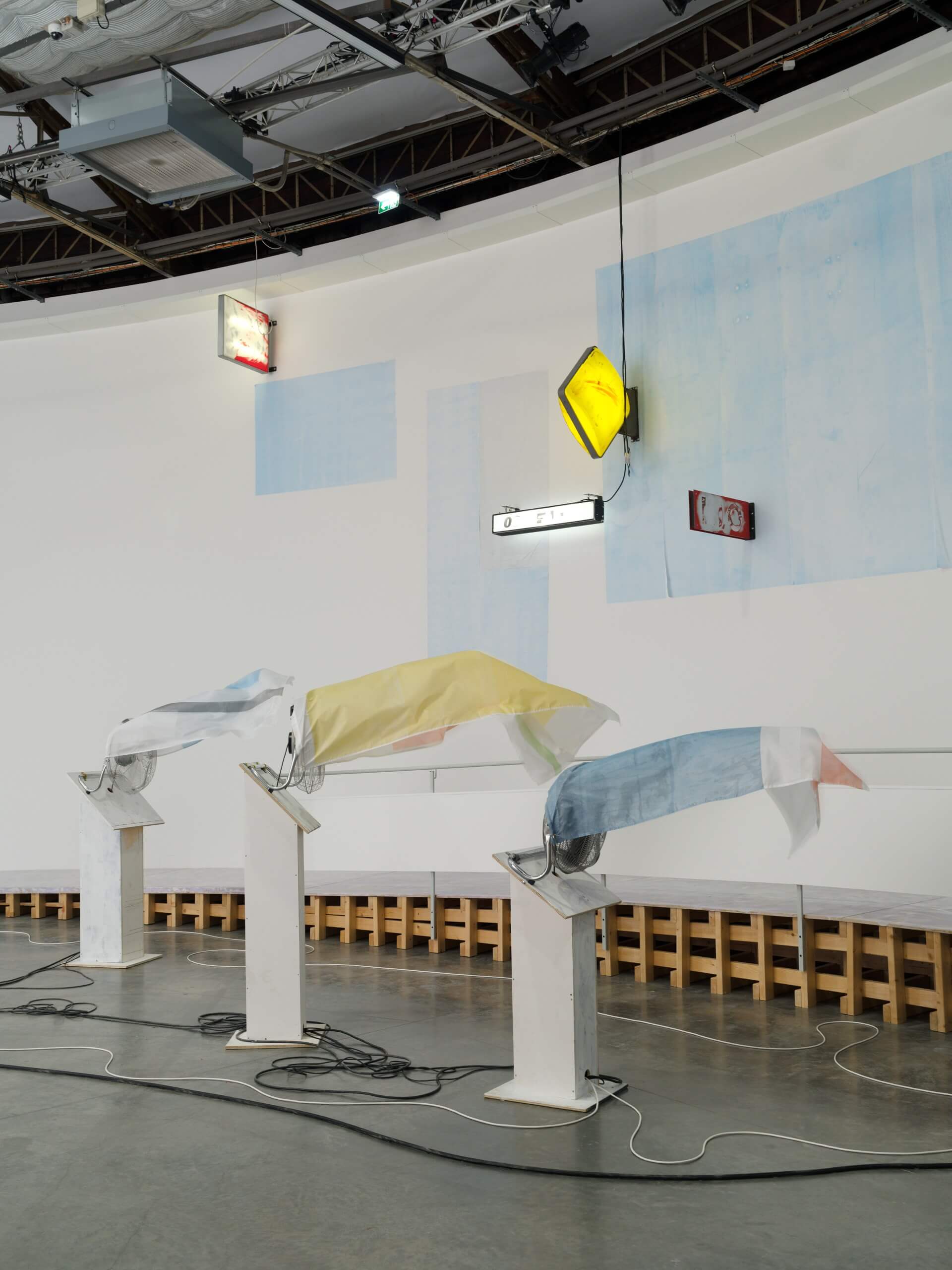 Three fabric sheets in yellow, blue, and white draped over white stands; illuminated signs are mounted on a curved white wall in the background.