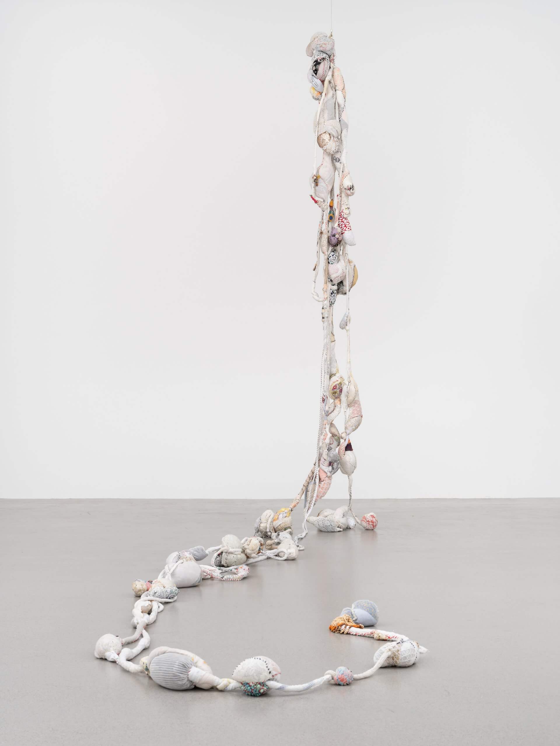 Dans un espace d'exposition minimaliste, une longue sculpture textile verticale, composée de faisceaux de tissu noués et enroulés, s'étend du plafond jusqu'au sol.
