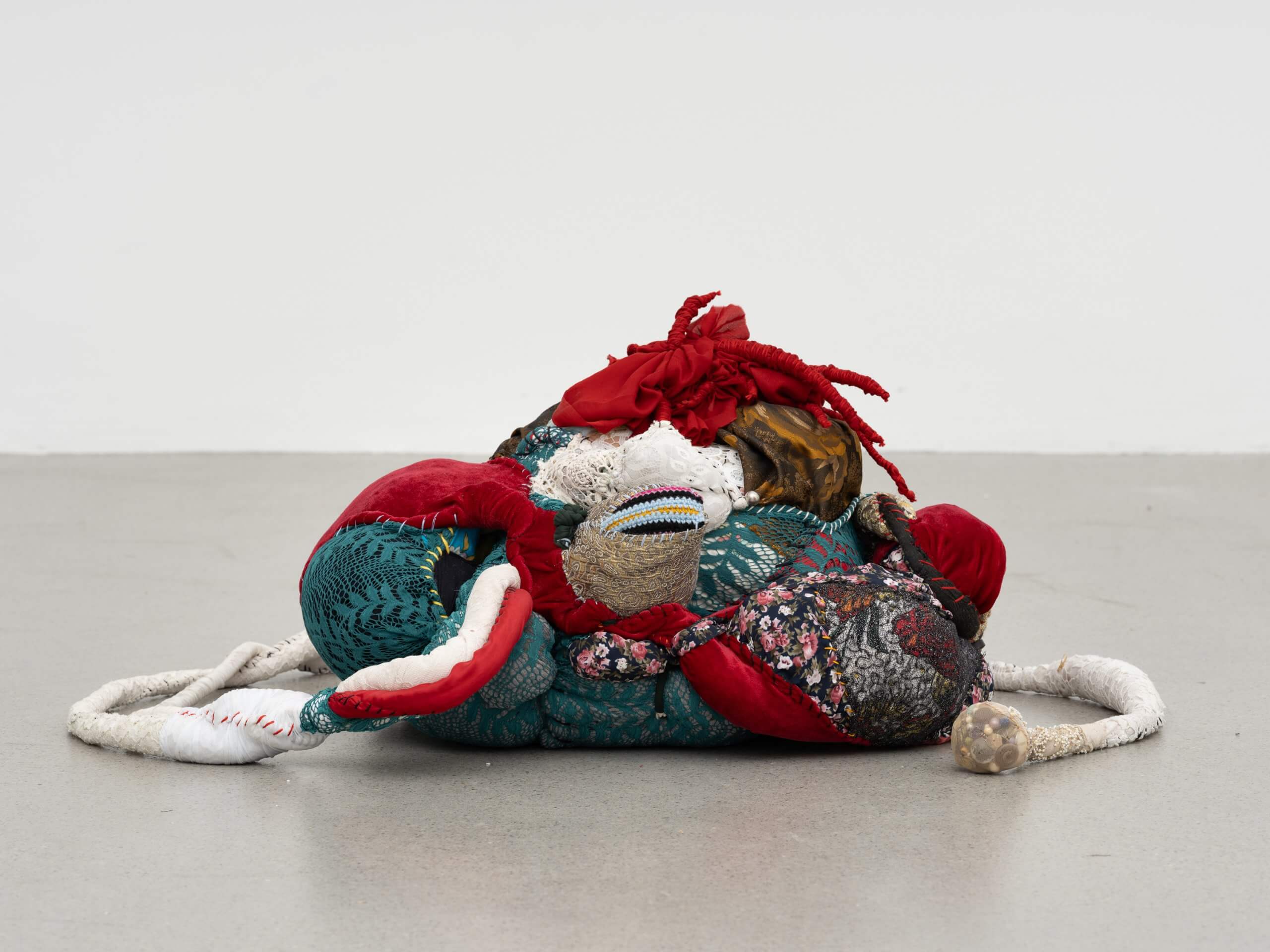 Une sculpture souple, colorée et abstraite, réalisée à partir de divers tissus aux motifs et textures variés, est disposée sur un sol gris uni, devant un fond blanc.