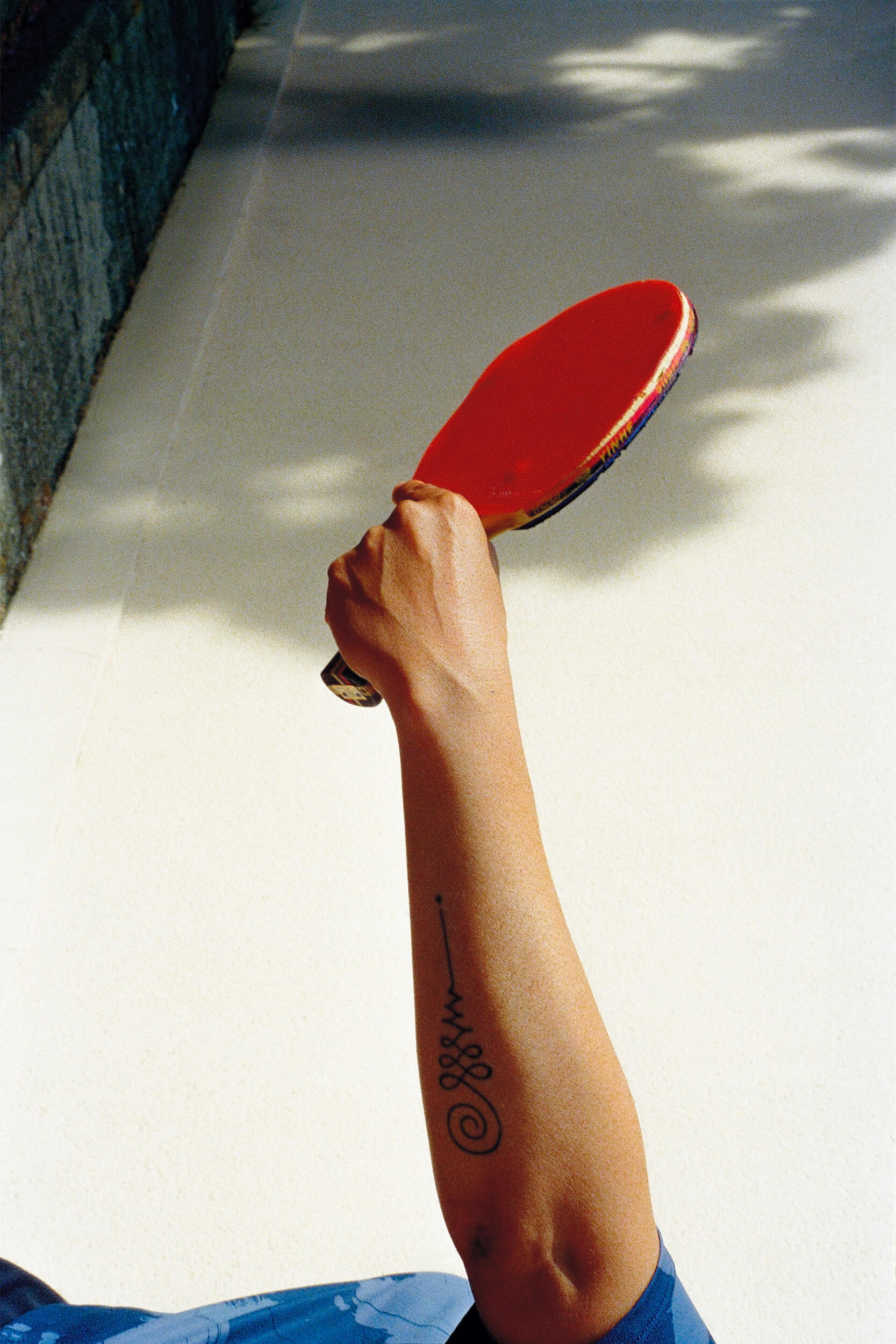 Une personne tenant une raquette de ping-pong rouge dans la main droite, avec un tatouage visible sur l'avant-bras, debout sur une surface ensoleillée.
