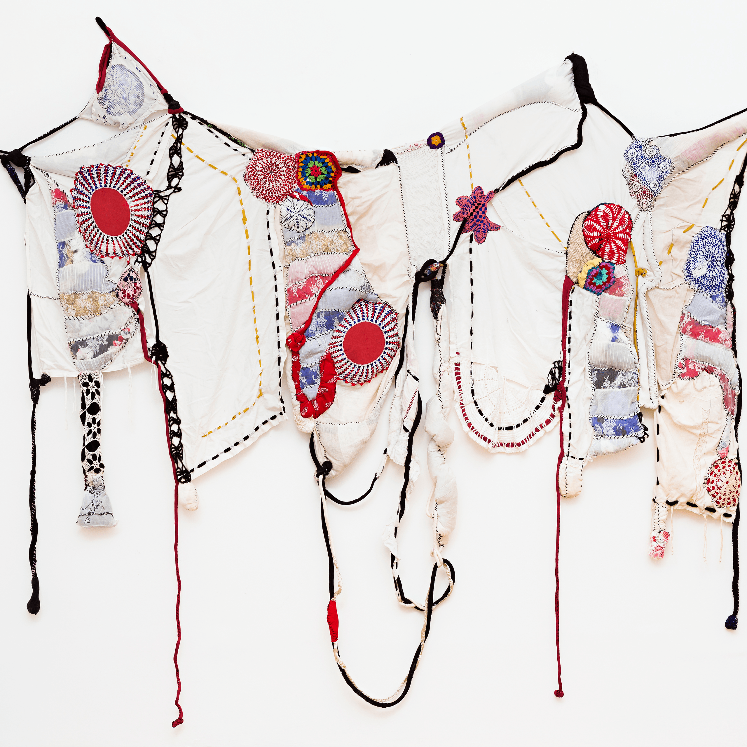 Cette tenture murale en tissu se compose de divers morceaux cousus, de motifs circulaires et de fils pendants de couleur rouge, bleue, noire et blanche sur fond blanc.