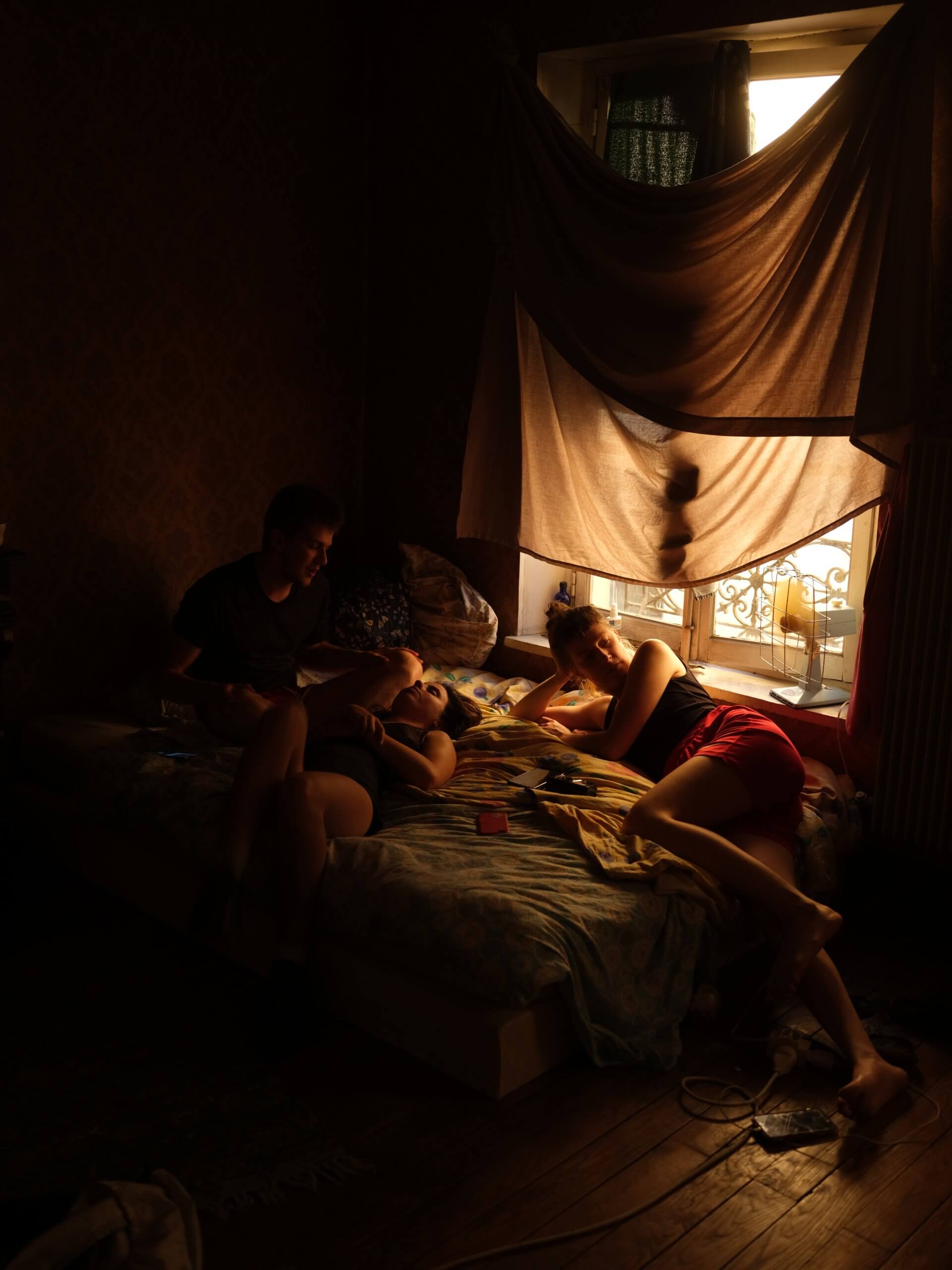 Trois personnes se détendent sur un lit dans une pièce faiblement éclairée, où la lumière du soleil pénètre à travers une fenêtre recouverte de draps drapés.