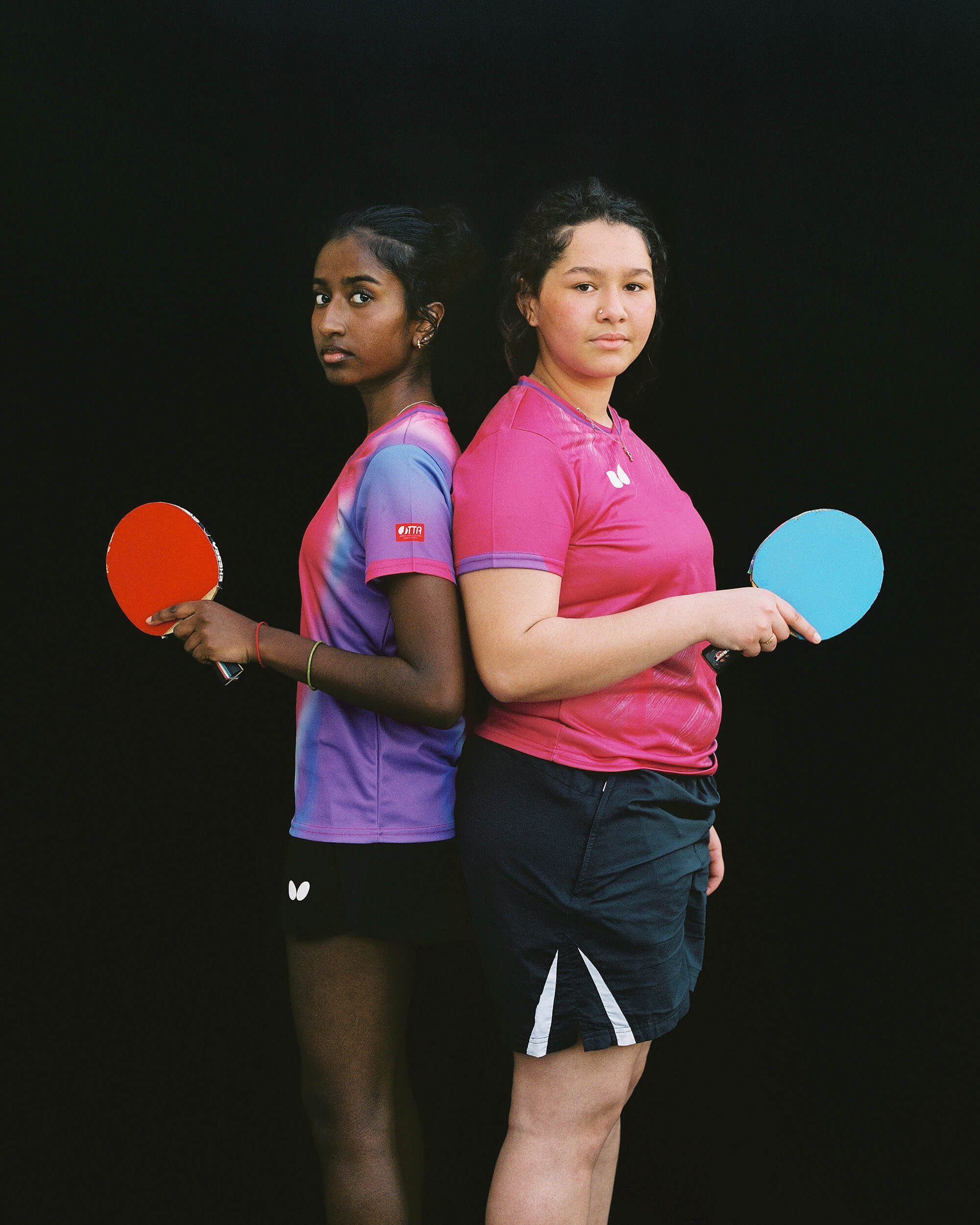 Deux jeunes femmes en tenue de sport se tiennent dos à dos, tenant chacune une raquette de tennis de table, l'une rouge et l'autre bleue, sur un fond noir uni.