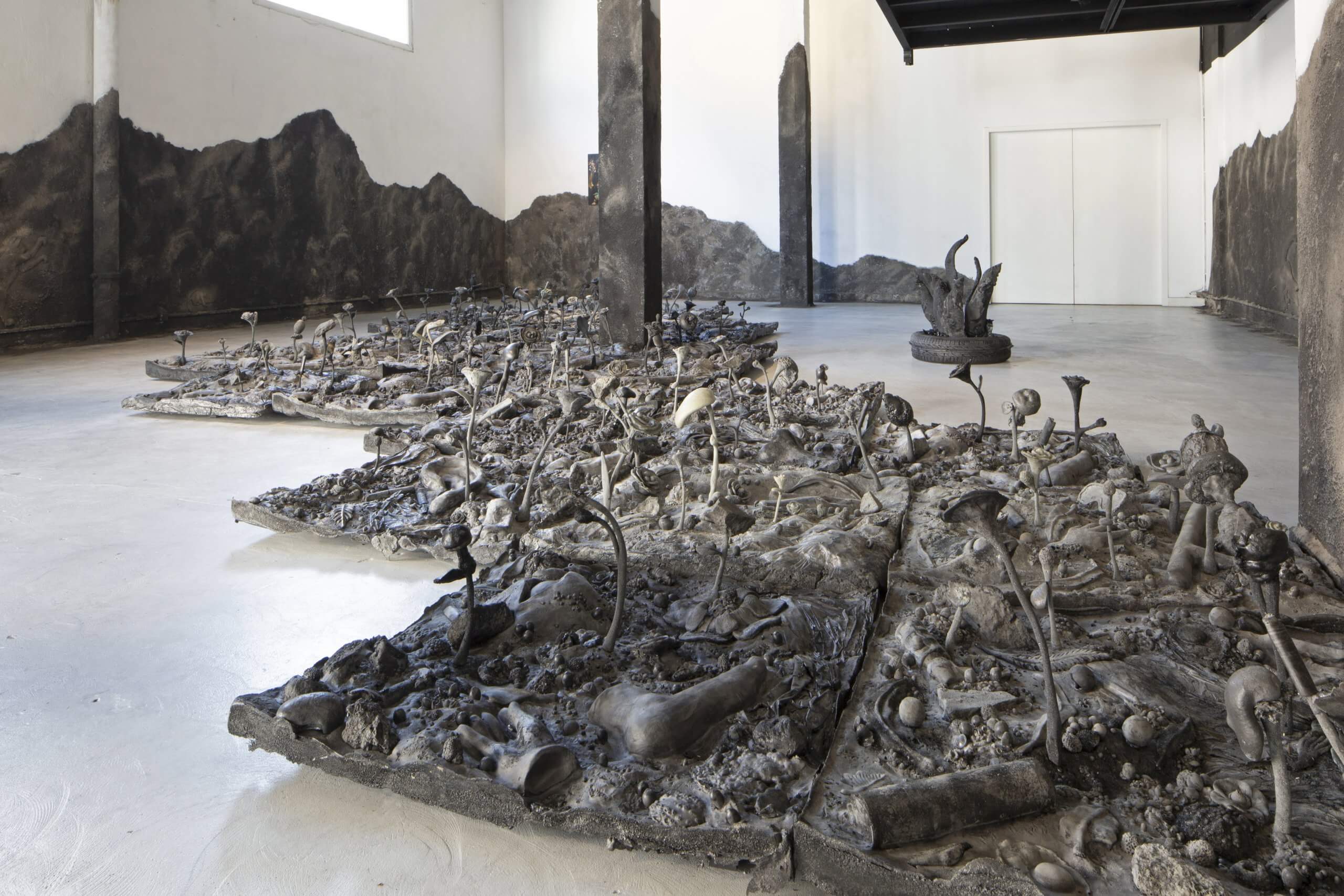 Grande installation sculpturale représentant des objets calcinés et fragmentés ainsi que des os, disposés sur des panneaux dans un espace d'exposition aux murs blancs, orné d'une fresque murale représentant des montagnes sombres.