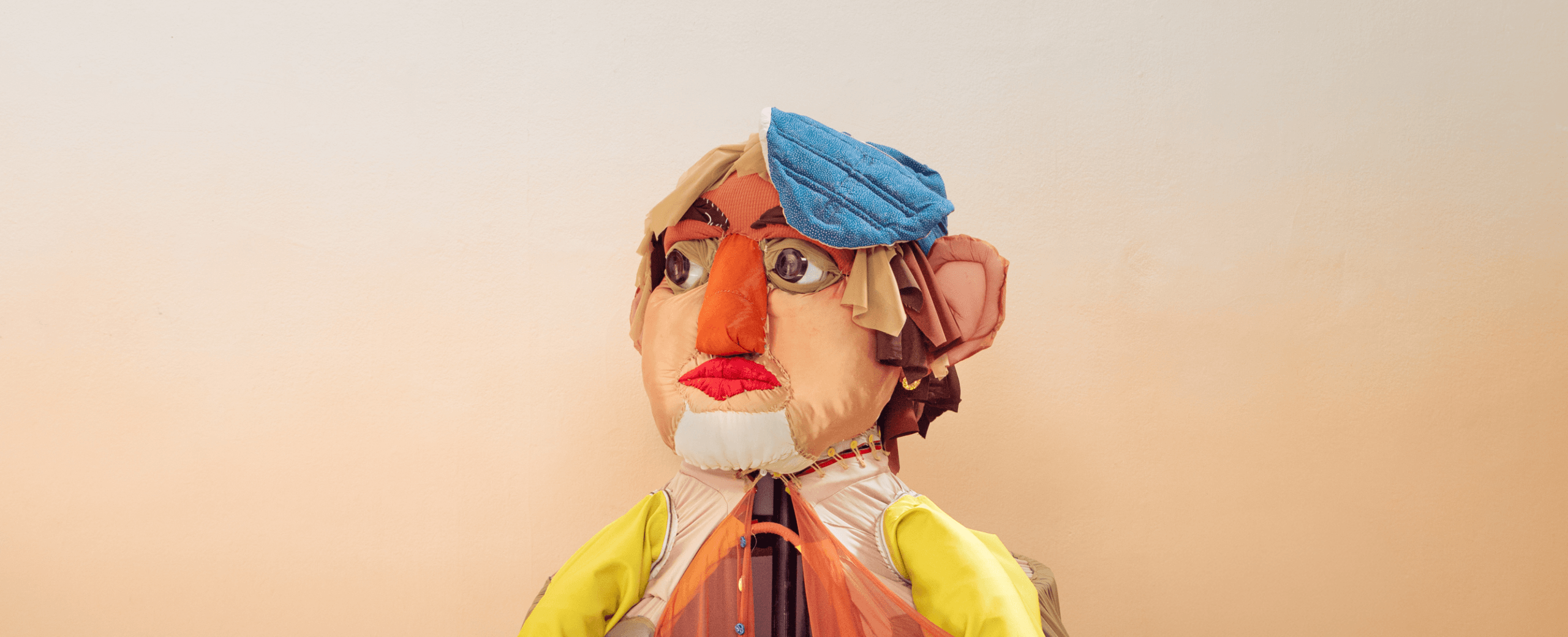 Une marionnette colorée aux traits exagérés, avec de grandes lèvres rouges, un cache-œil bleu sur la tête et des gants en caoutchouc en guise de bras, posant devant un fond beige.