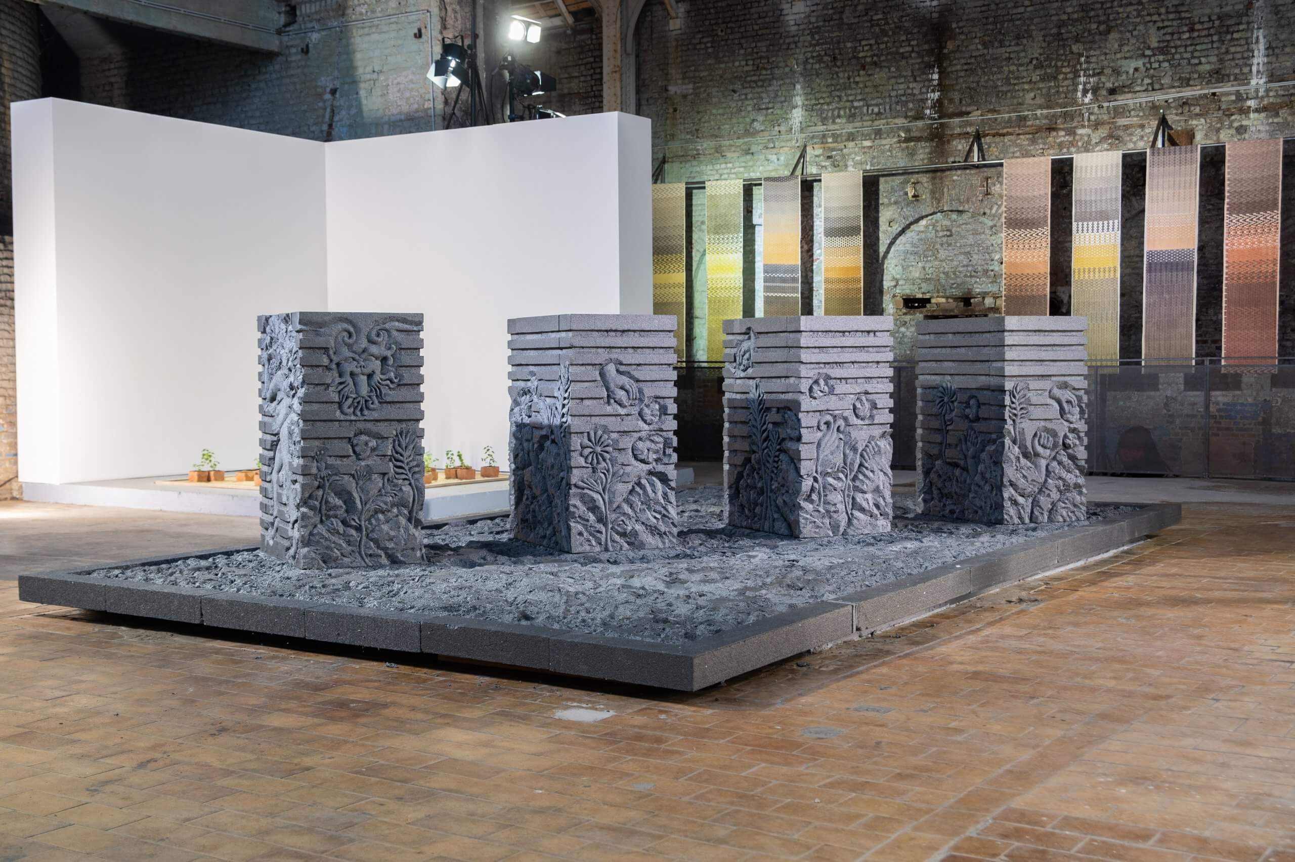 Quatre piliers en pierre grise ornés de motifs sculptés se dressent sur une estrade rectangulaire, dans un espace d'exposition intérieur aux murs en briques où sont présentées des œuvres d'art.