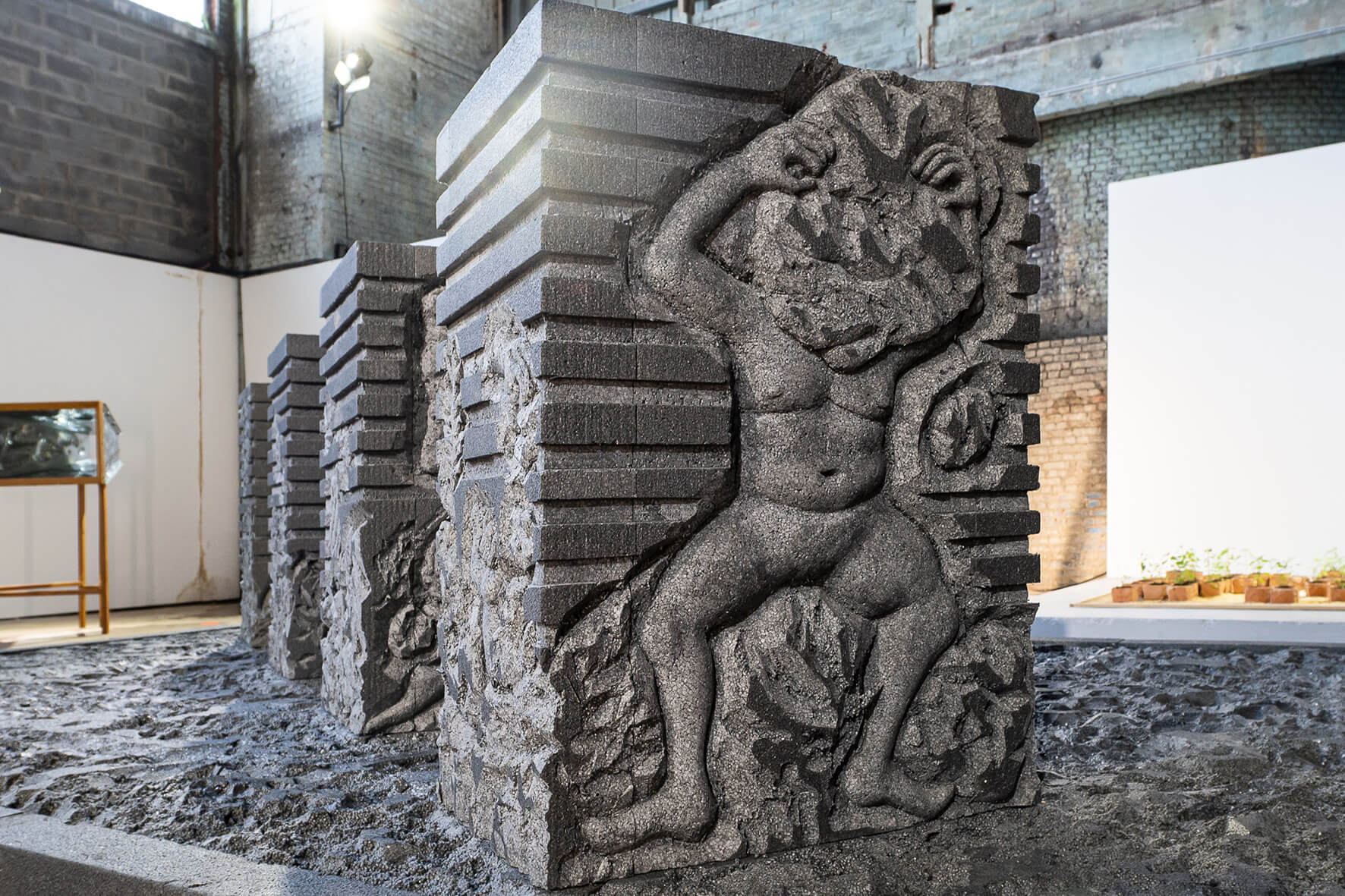 Une sculpture en pierre taillée représente une silhouette humaine émergeant de blocs de pierre abstraits et superposés, dans un espace d'exposition intérieur.