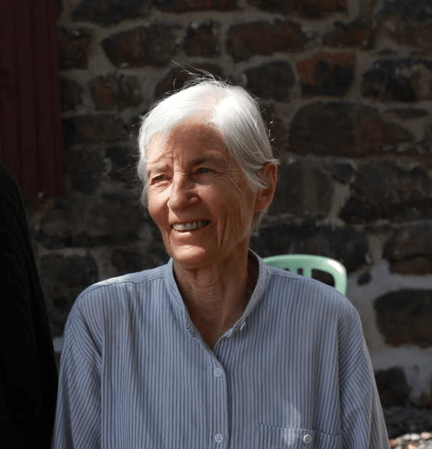 Une femme d'un certain âge, aux cheveux blancs courts et vêtue d'un t-shirt rayé, sourit debout à l'extérieur, devant un mur de pierre.