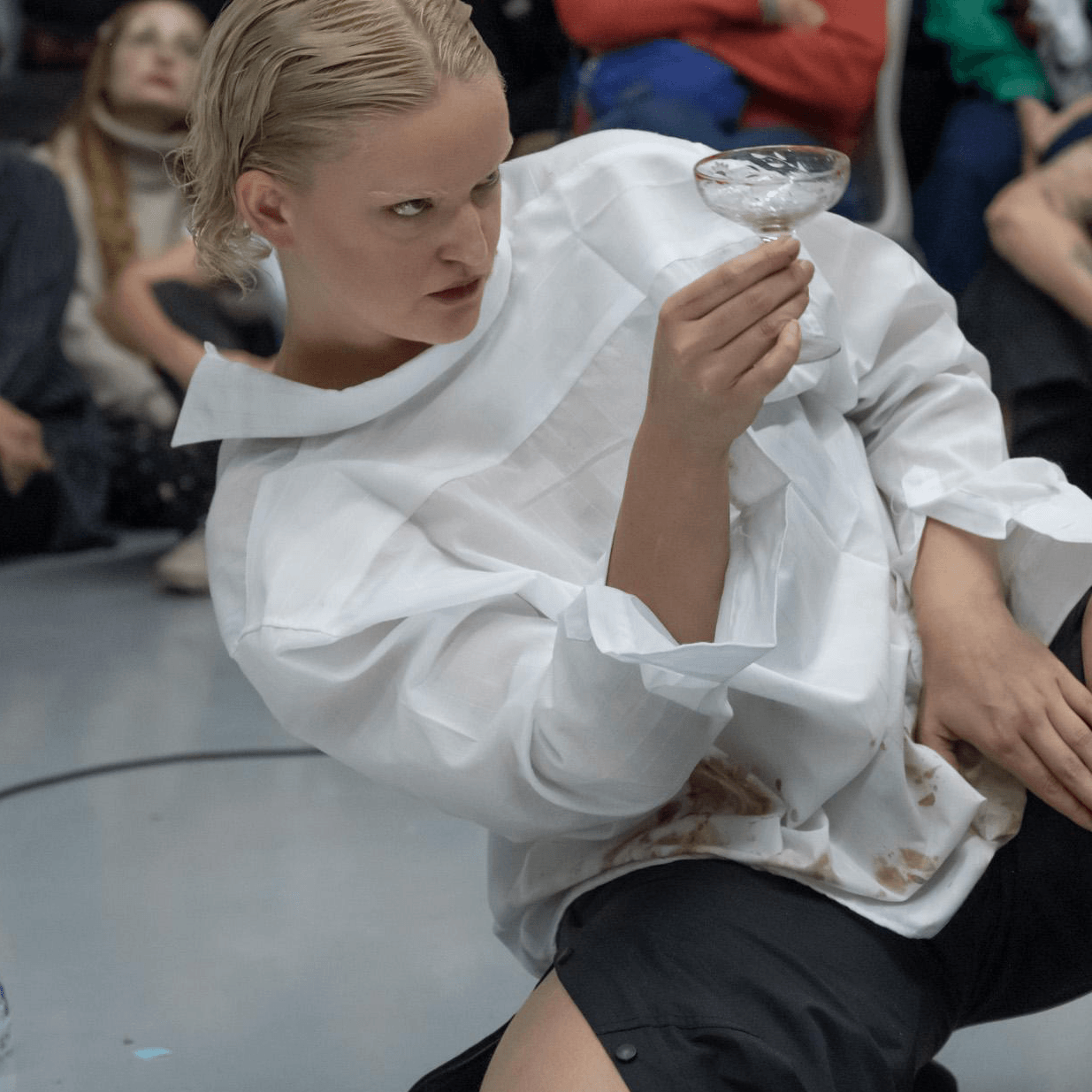 Une personne vêtue d'une chemise blanche et d'un pantalon noir est assise par terre, tenant un verre à pied qu'elle regarde attentivement. Des personnes sont assises à l'arrière-plan.