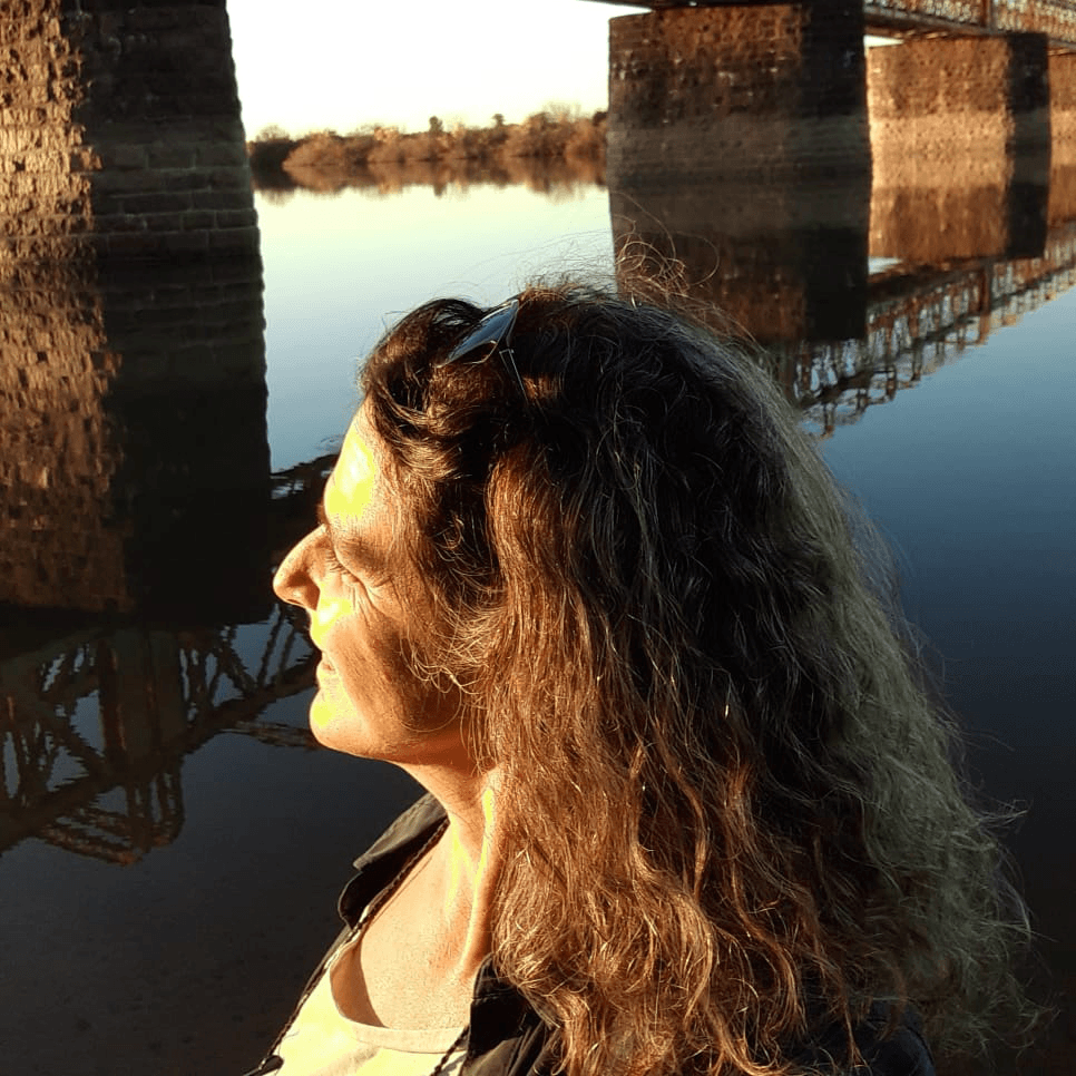 Une femme aux cheveux bouclés se tient près d'une eau calme au coucher du soleil, avec un pont et son reflet visible à l'arrière-plan.