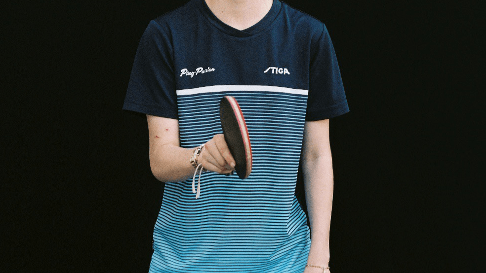 Une personne vêtue d'un t-shirt rayé bleu, tenant une raquette de tennis de table, sur un fond noir uni.