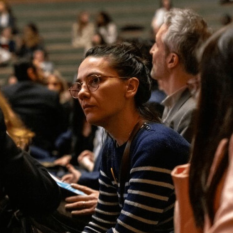 Une femme portant des lunettes et un pull rayé est assise parmi un groupe de personnes dans ce qui semble être une salle de spectacle.