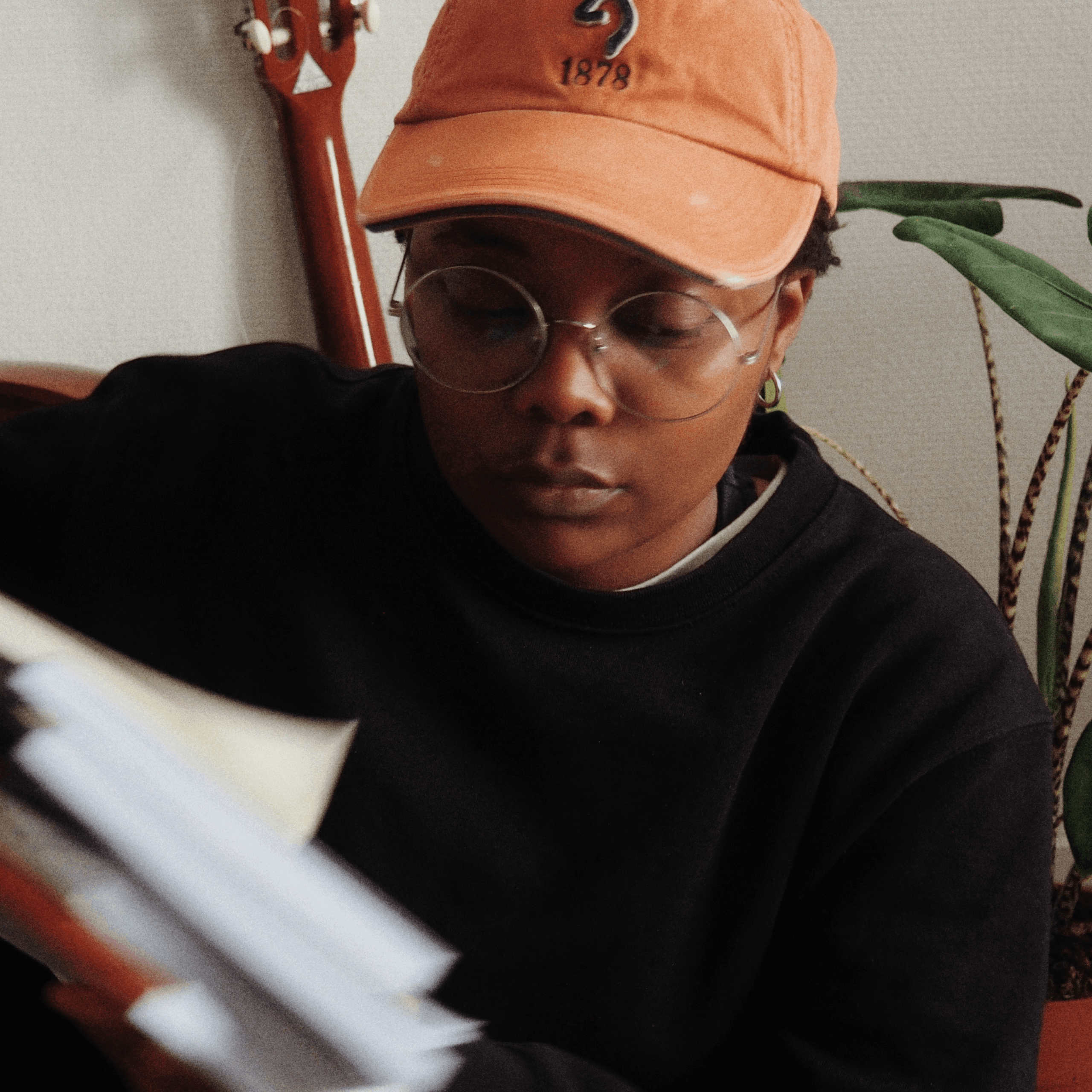 Une personne portant des lunettes et une casquette orange lit ou organise une pile de papiers, assise à l'intérieur près d'une plante et d'une guitare.