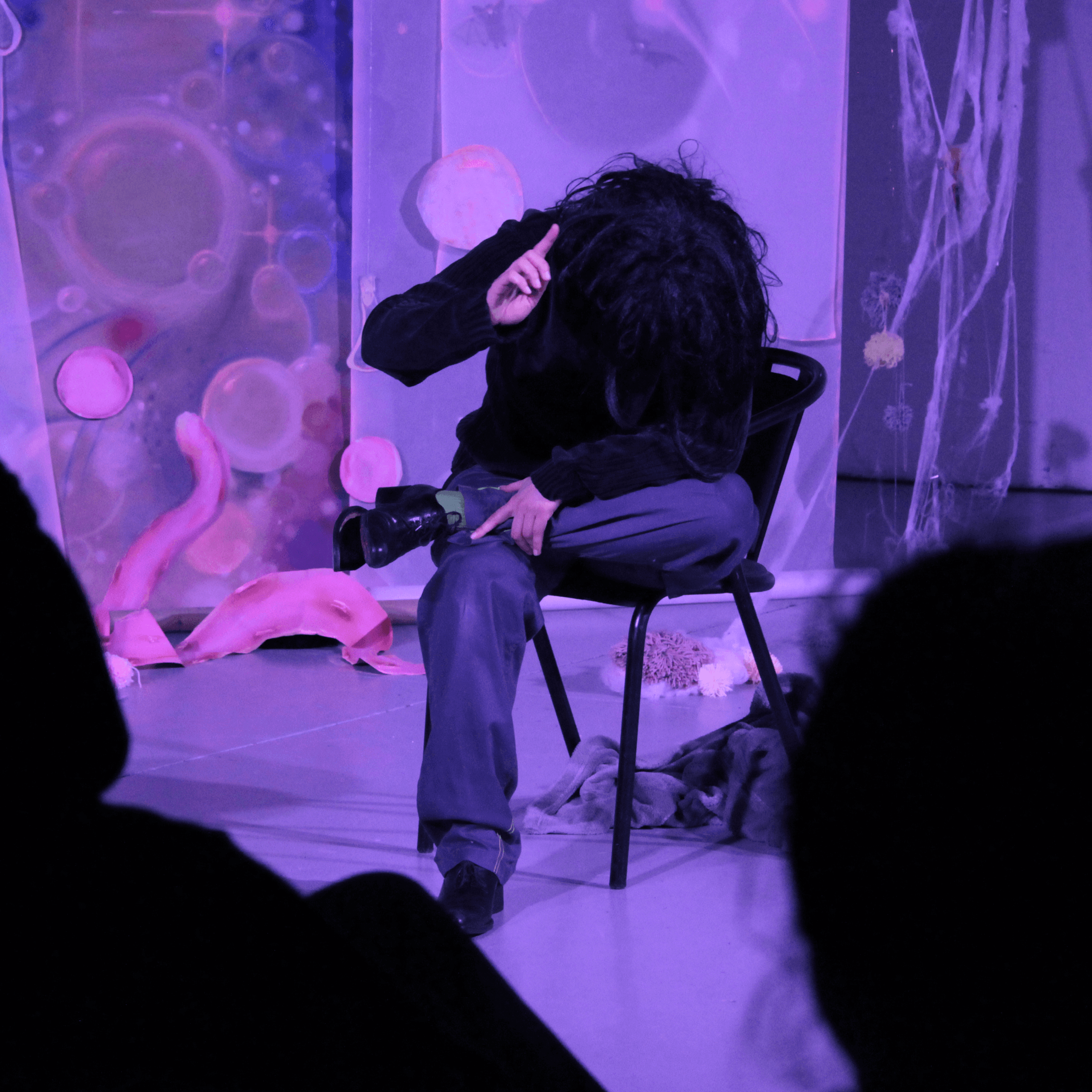 Une personne aux longs cheveux noirs est assise sur une chaise, la tête baissée et une main levée, dans une pièce éclairée en violet avec des décorations abstraites et des silhouettes de spectateurs assis au premier plan.