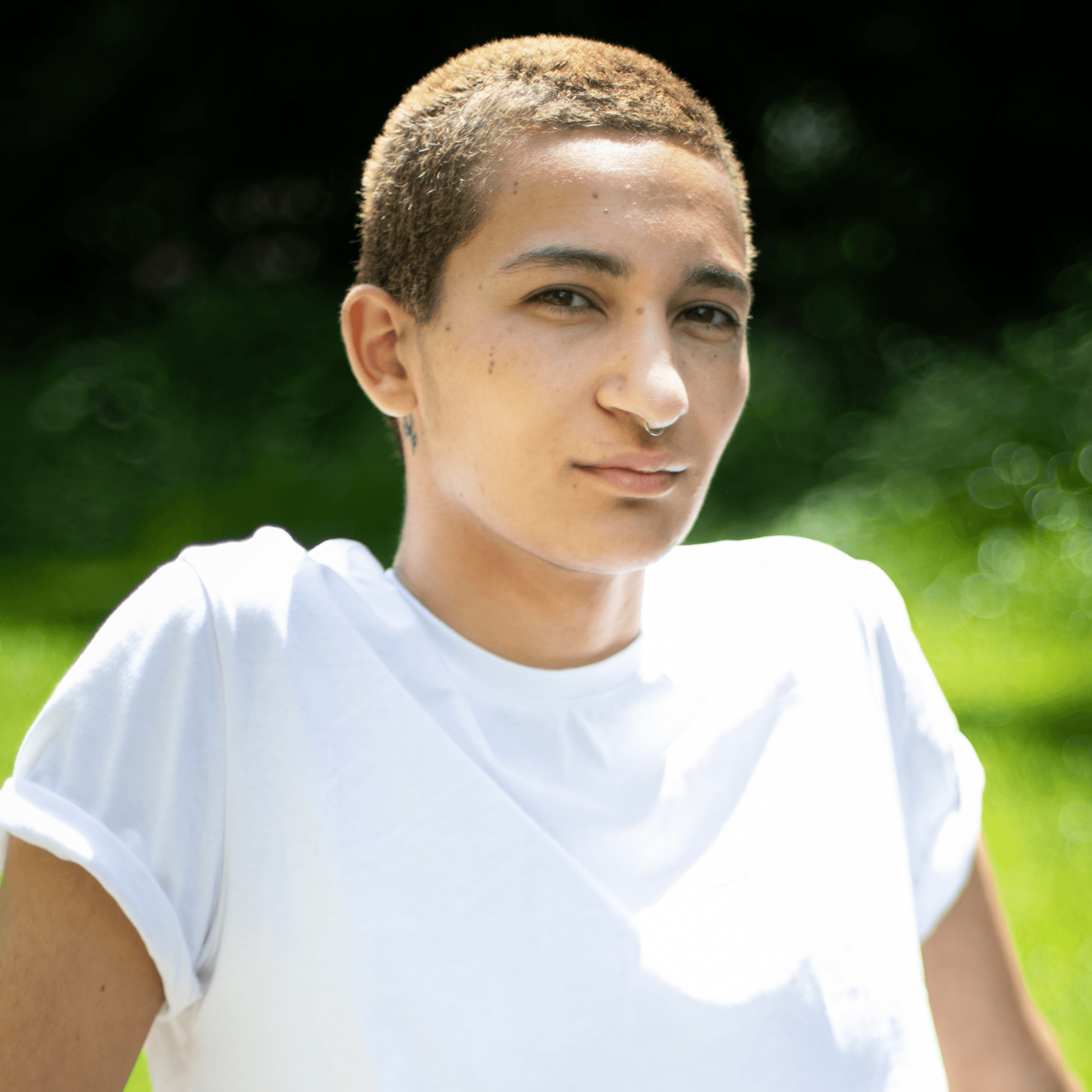 Une personne aux cheveux courts et châtains clairs portant un t-shirt blanc se tient à l'extérieur, en plein soleil, avec de la verdure floue en arrière-plan.