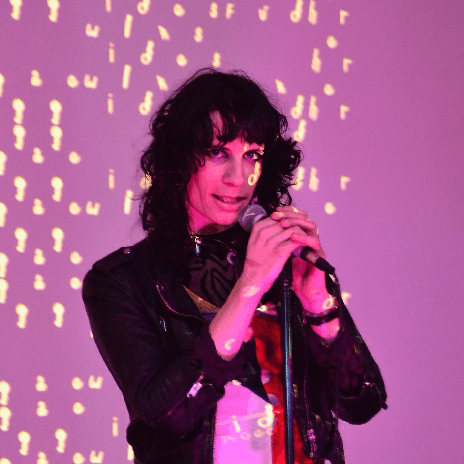 Personne aux longs cheveux noirs tenant un microphone, debout devant un fond violet avec des motifs lumineux jaunes dispersés.