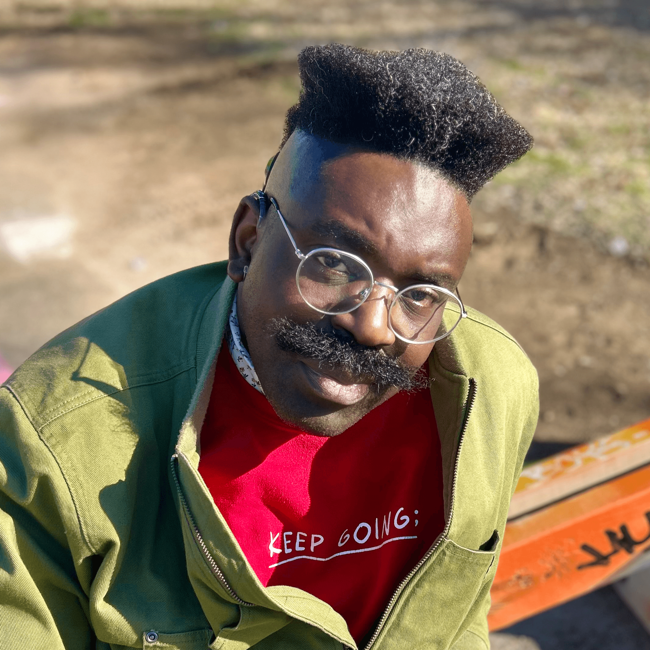 Une personne avec une coiffure décolorée, des lunettes et une moustache porte une chemise rouge sur laquelle on peut lire KEEP GOING et une veste verte, assise à l'extérieur par une journée ensoleillée.