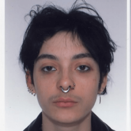 Personne aux cheveux courts et foncés, portant un haut sombre, regardant directement l'appareil photo sur un fond uni, avec plusieurs piercings aux oreilles et au septum.