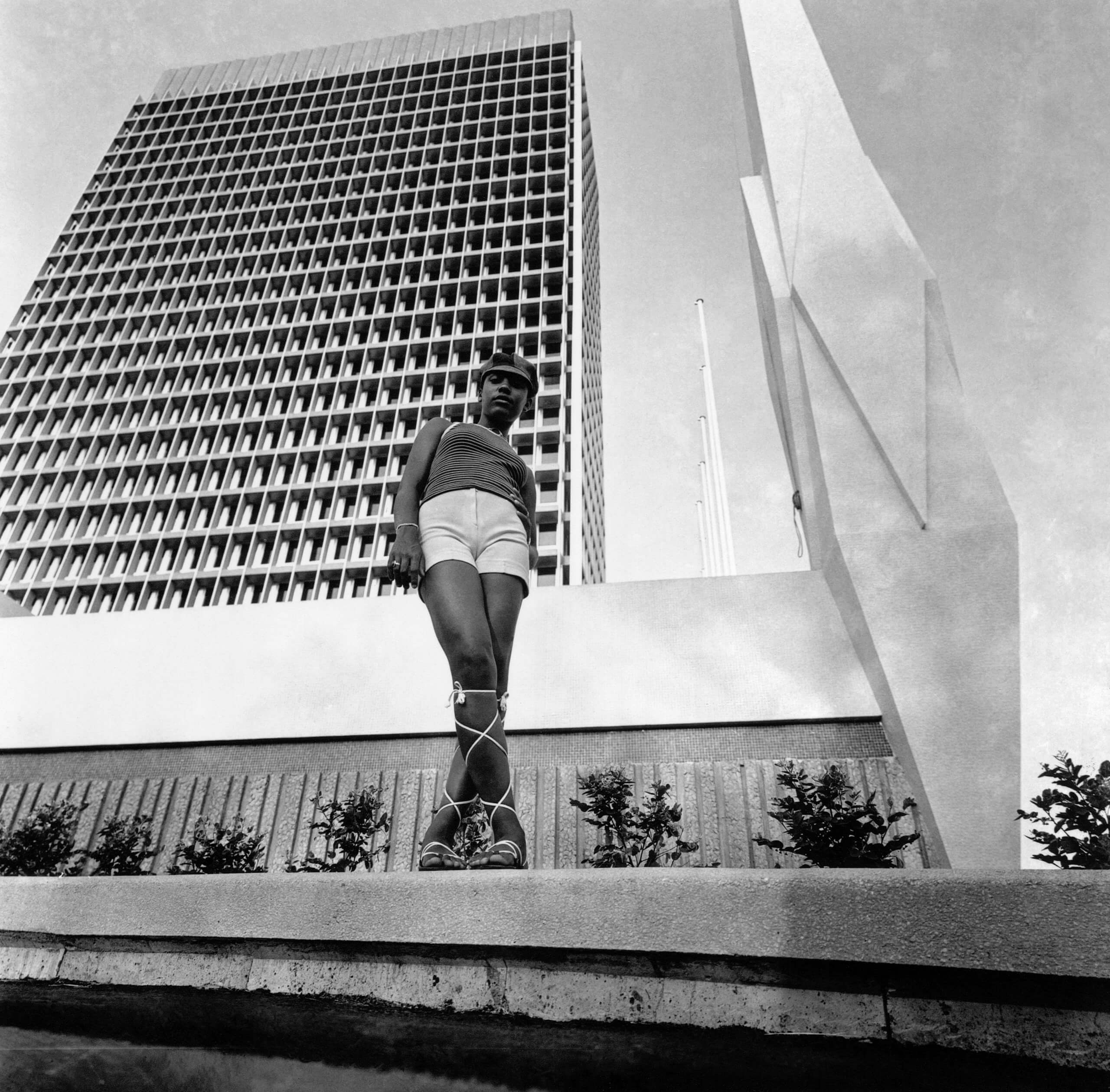 Une personne portant un chapeau, un short et des sandales se tient près d'une sculpture moderne et d'un grand immeuble de bureaux, avec des arbres et un muret en arrière-plan.