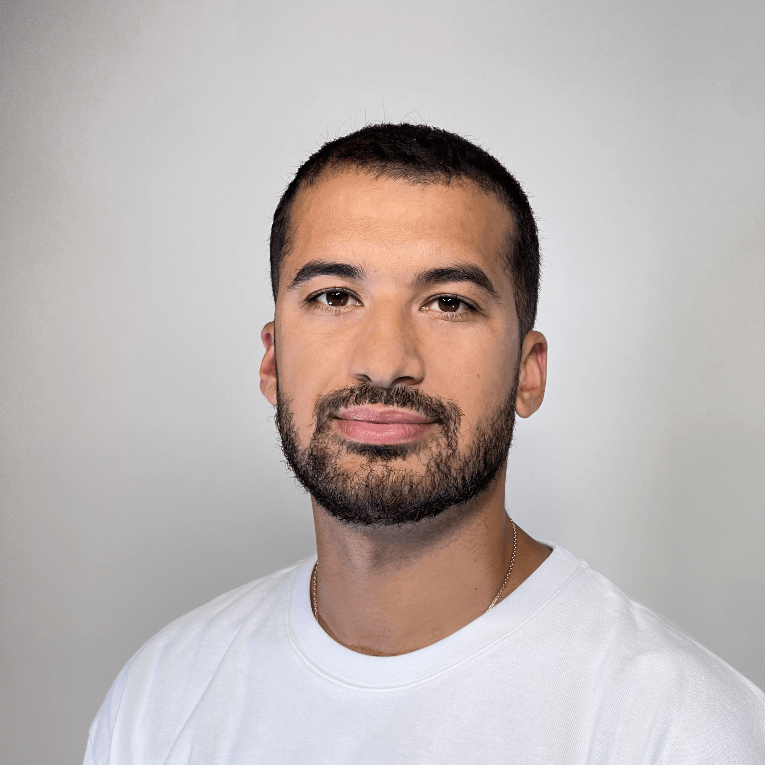 Un homme aux cheveux courts et foncés, portant une barbe, un t-shirt blanc et un collier en or, pose devant un fond clair et uni.