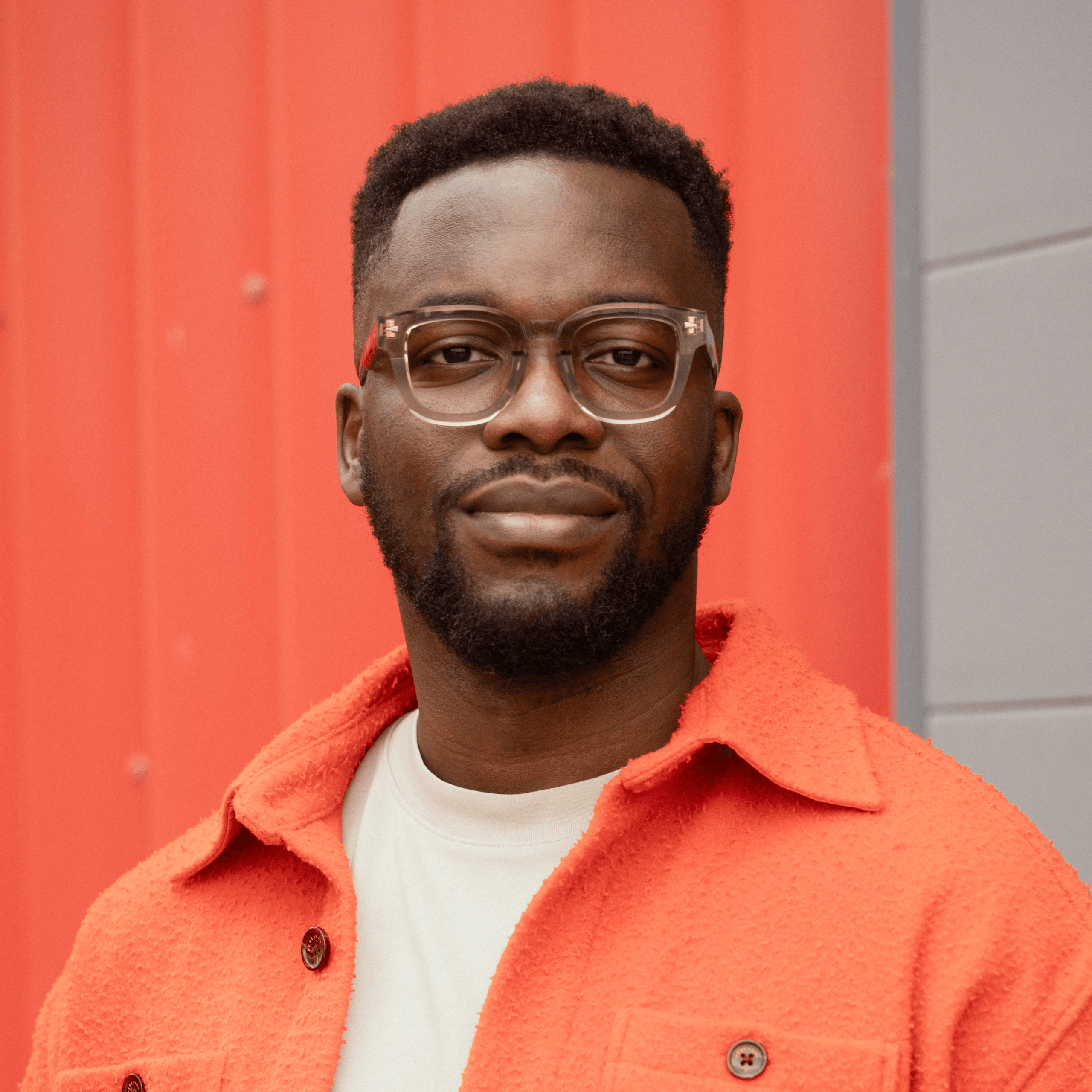 Un homme aux cheveux courts et à la barbe porte des lunettes et une veste orange par-dessus une chemise blanche ; il se tient debout devant un mur rouge et gris.