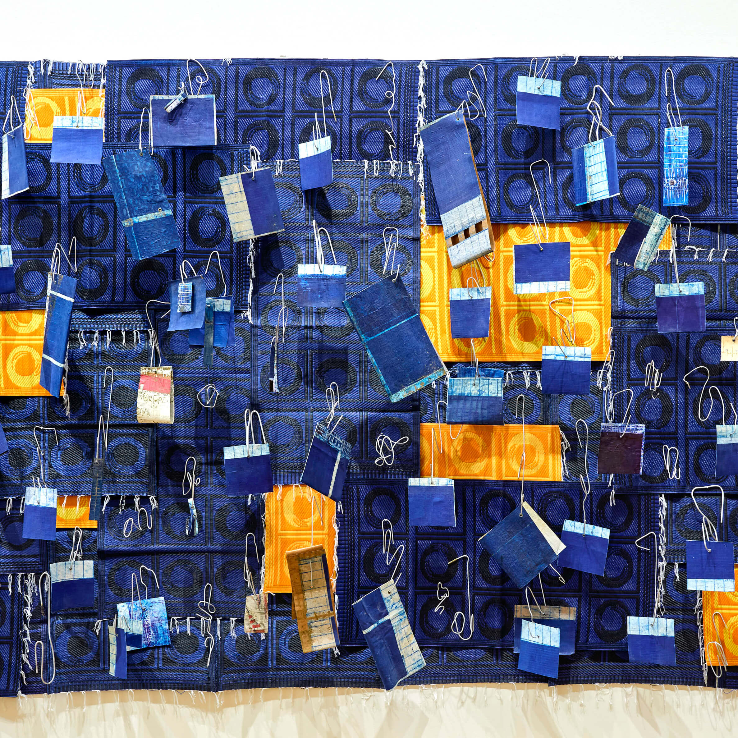 Œuvre d'art textile composée d'un tissu à motifs géométriques bleus et jaunes, sur lequel sont fixés de petits morceaux de tissu rectangulaires bleus et multicolores, maintenus par des ficelles blanches.