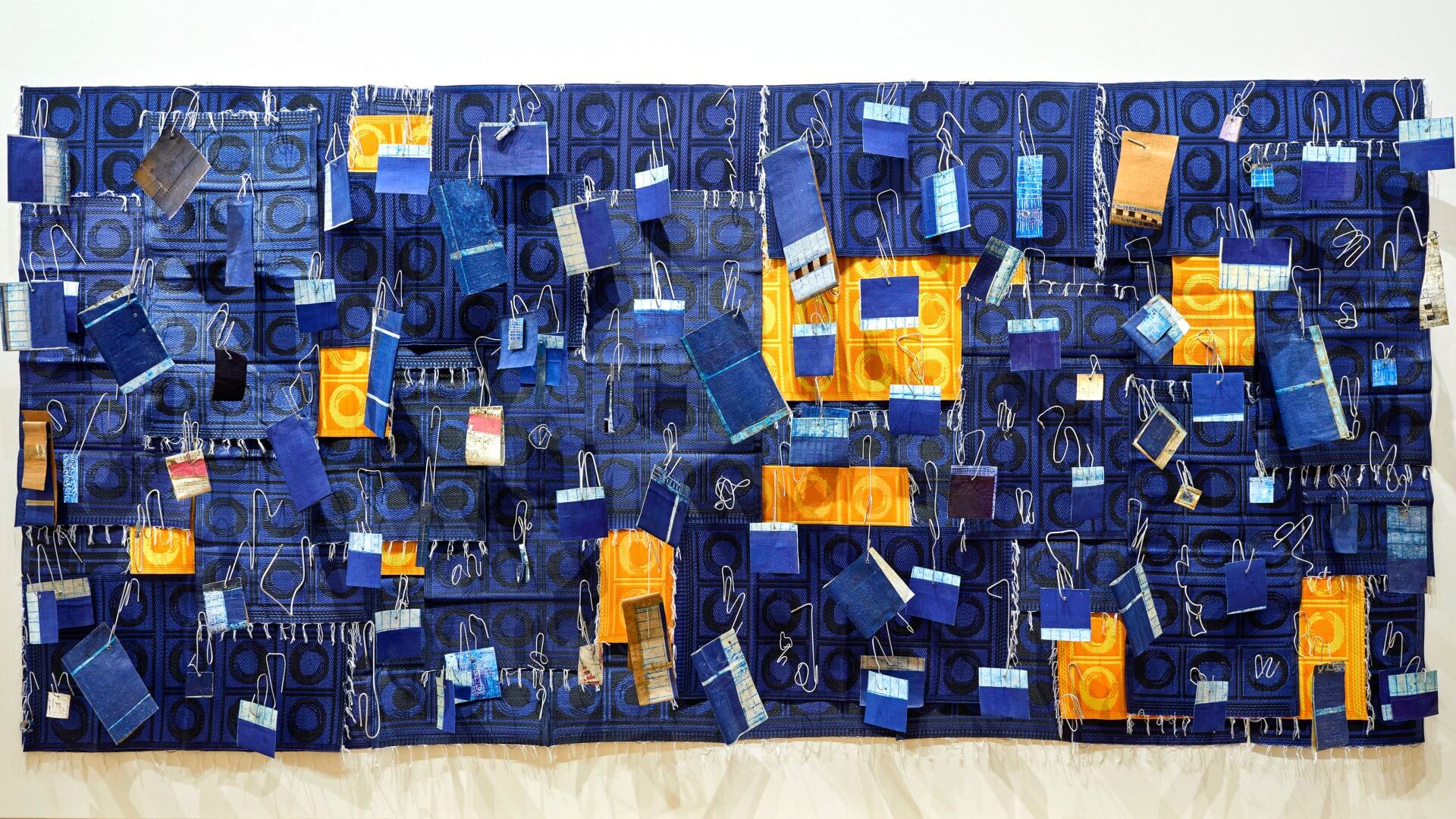 Une grande œuvre d'art textile composée de morceaux de tissu bleus et jaunes, de crochets métalliques et d'échantillons disposés sur un fond bleu à motifs.