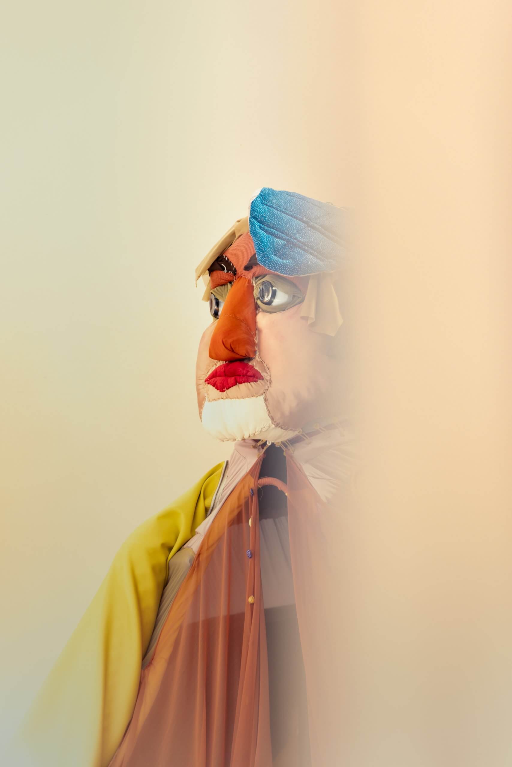 Une grande marionnette colorée aux traits exagérés, coiffée d'une coiffe bleue et vêtue d'une tenue jaune et orange, partiellement dissimulée par un voile beige flou.