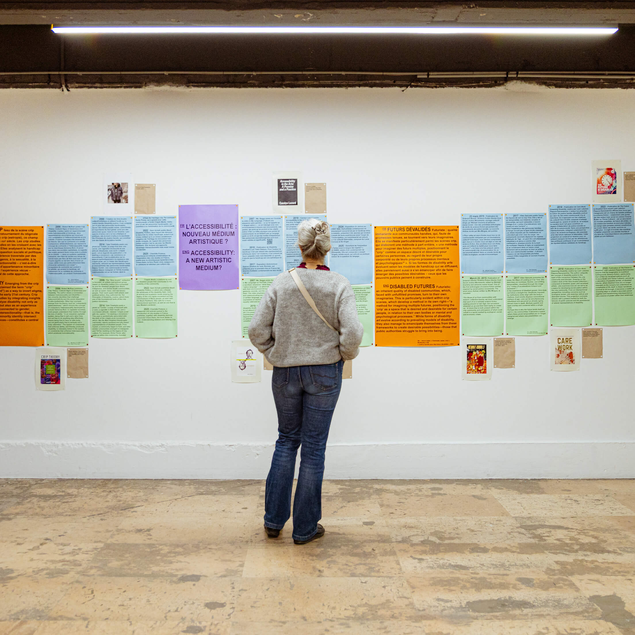 Une personne se tient devant un mur blanc recouvert d'affiches imprimées aux couleurs vives et de textes, dans le cadre d'une exposition ou d'une galerie.