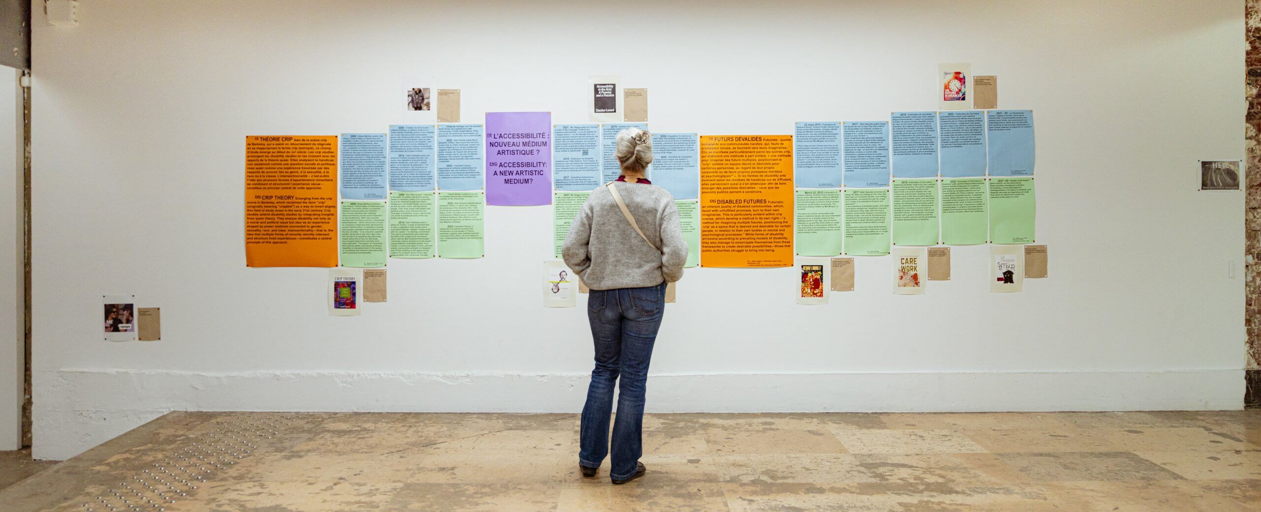 Une personne se tient devant un mur de galerie sur lequel sont accrochés des affiches colorées et des documents écrits, disposés en rangées.