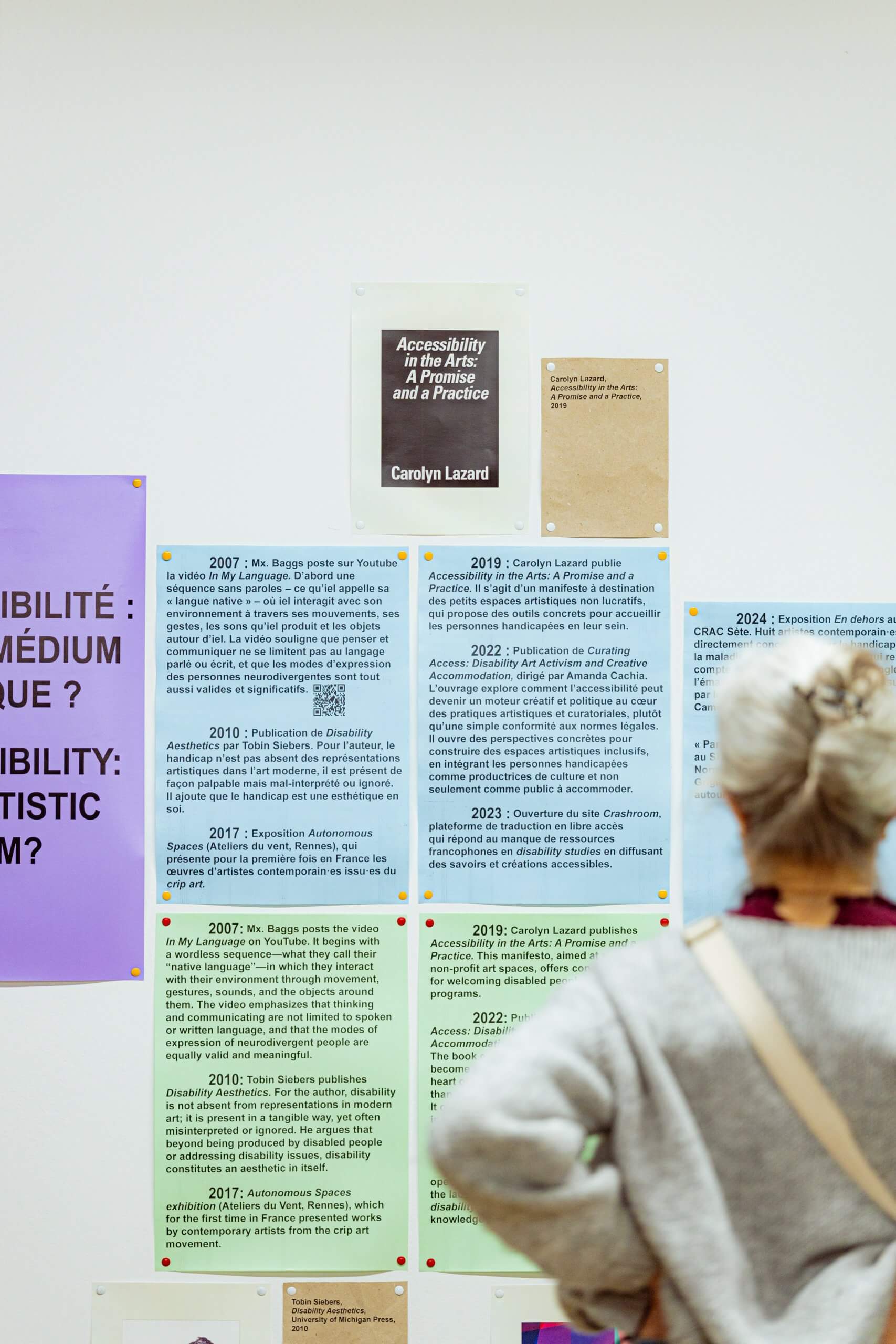 Une personne aux cheveux gris, les cheveux relevés en chignon, observe une exposition murale composée d'affiches et de textes consacrés à l'accessibilité dans le domaine des arts, parmi lesquels figure un ouvrage de Carolyn Lazard mis en avant.