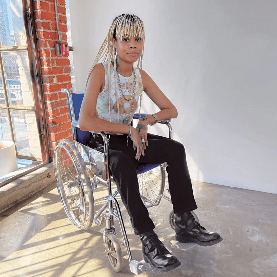 Une personne aux longues tresses claires est assise dans un fauteuil roulant près d'une fenêtre ensoleillée. Elle porte un haut sans manches, un pantalon noir et des bottes noires.