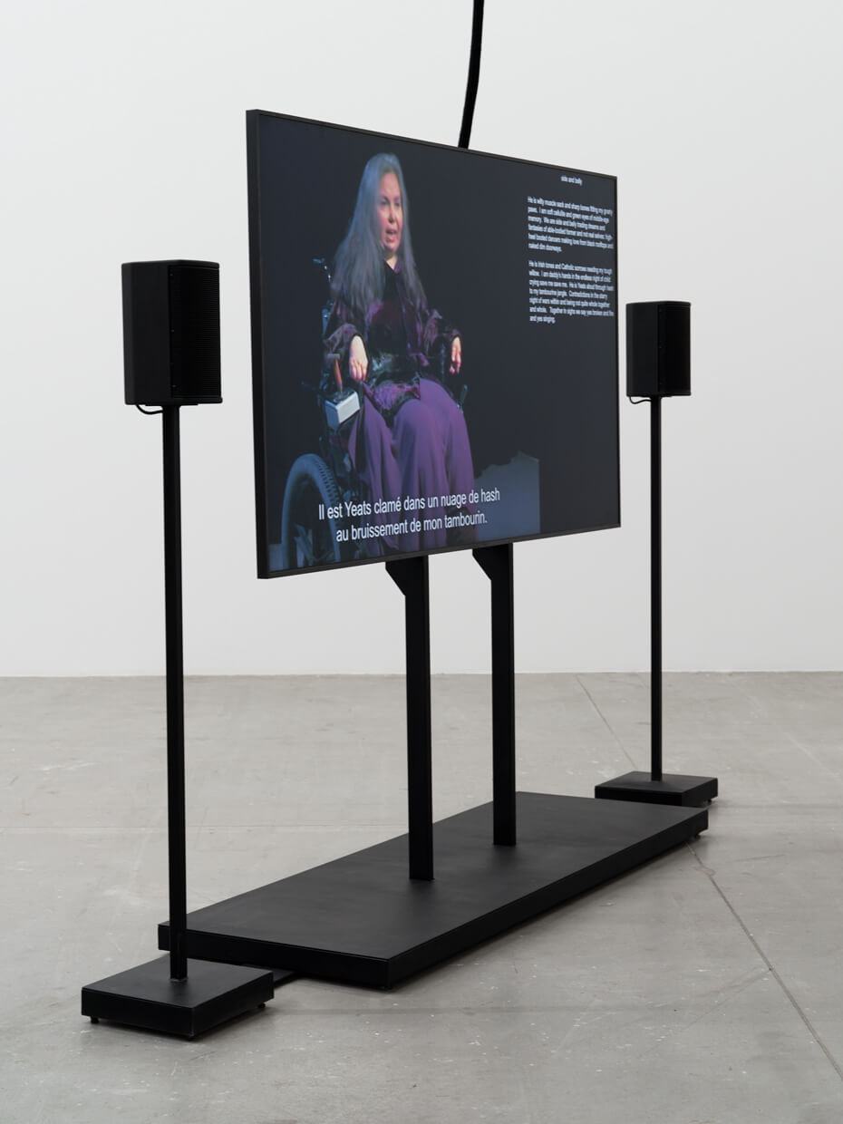 Une installation vidéo met en scène une femme assise qui s'exprime à l'écran, avec des sous-titres en français, flanquée de deux haut-parleurs noirs posés sur une estrade, dans un espace d'exposition au style minimaliste.