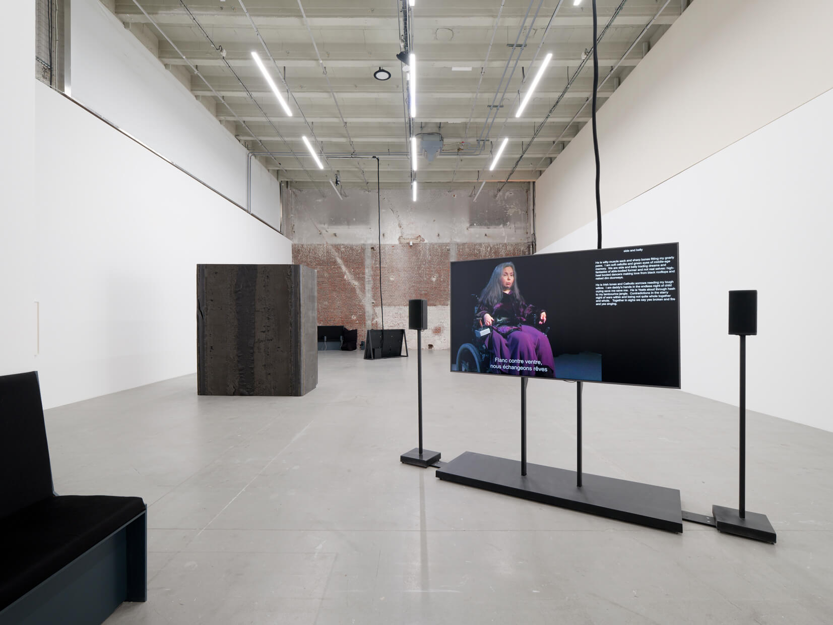 Une galerie d'art contemporain avec un écran de télévision sur lequel apparaît une femme s'exprimant en langue des signes, une sculpture cubique noire, un banc et un mur en briques apparentes en arrière-plan.