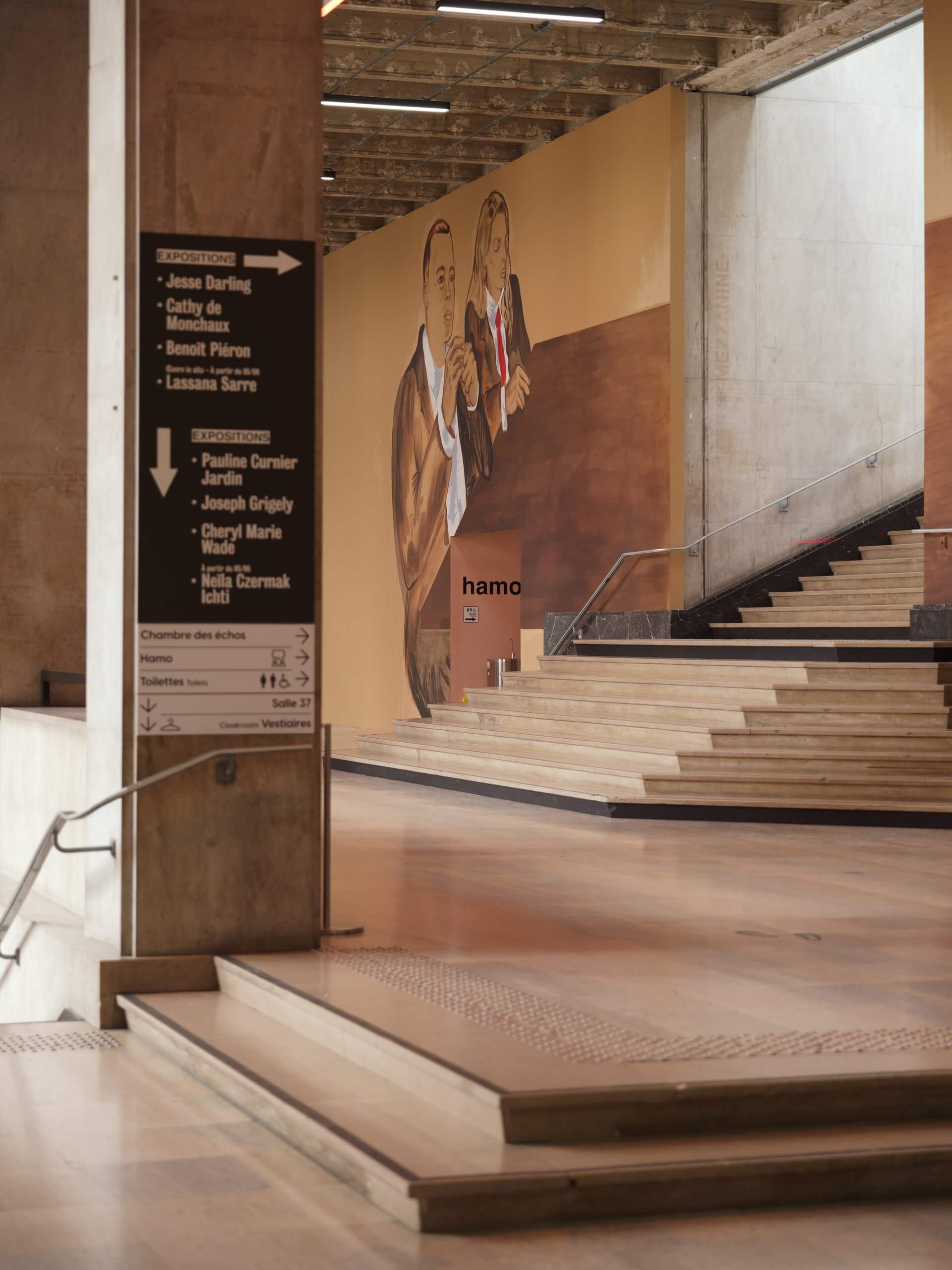 Un espace intérieur comportant des colonnes en béton, un escalier, un panneau indicateur et une grande fresque murale représentant deux personnes tenant des verres à la main, sur un mur aux tons beige et marron.