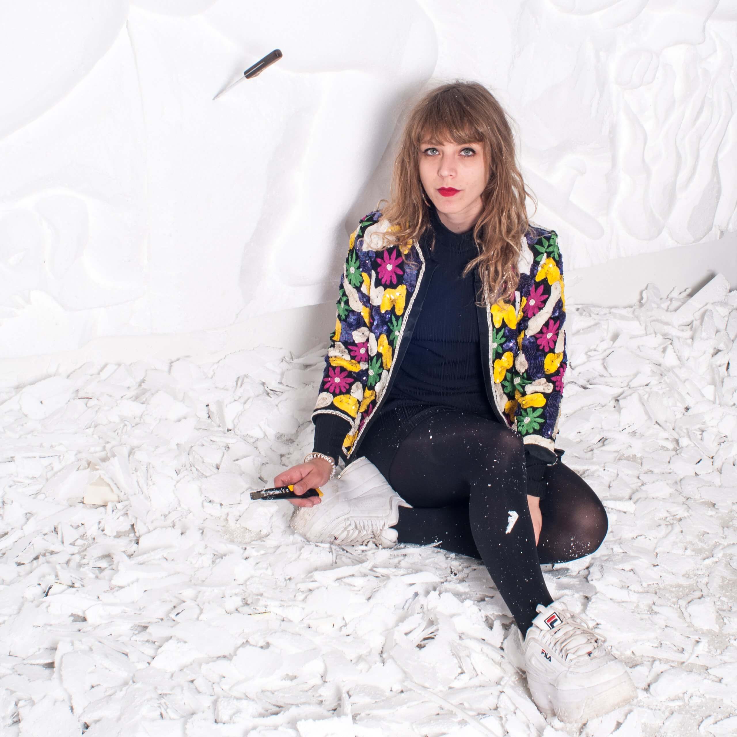 Une femme vêtue d'une veste à fleurs aux couleurs vives est assise sur un sol jonché de débris blancs ; elle tient un petit objet dans la main, tandis qu'un couteau est planté dans le mur derrière elle.