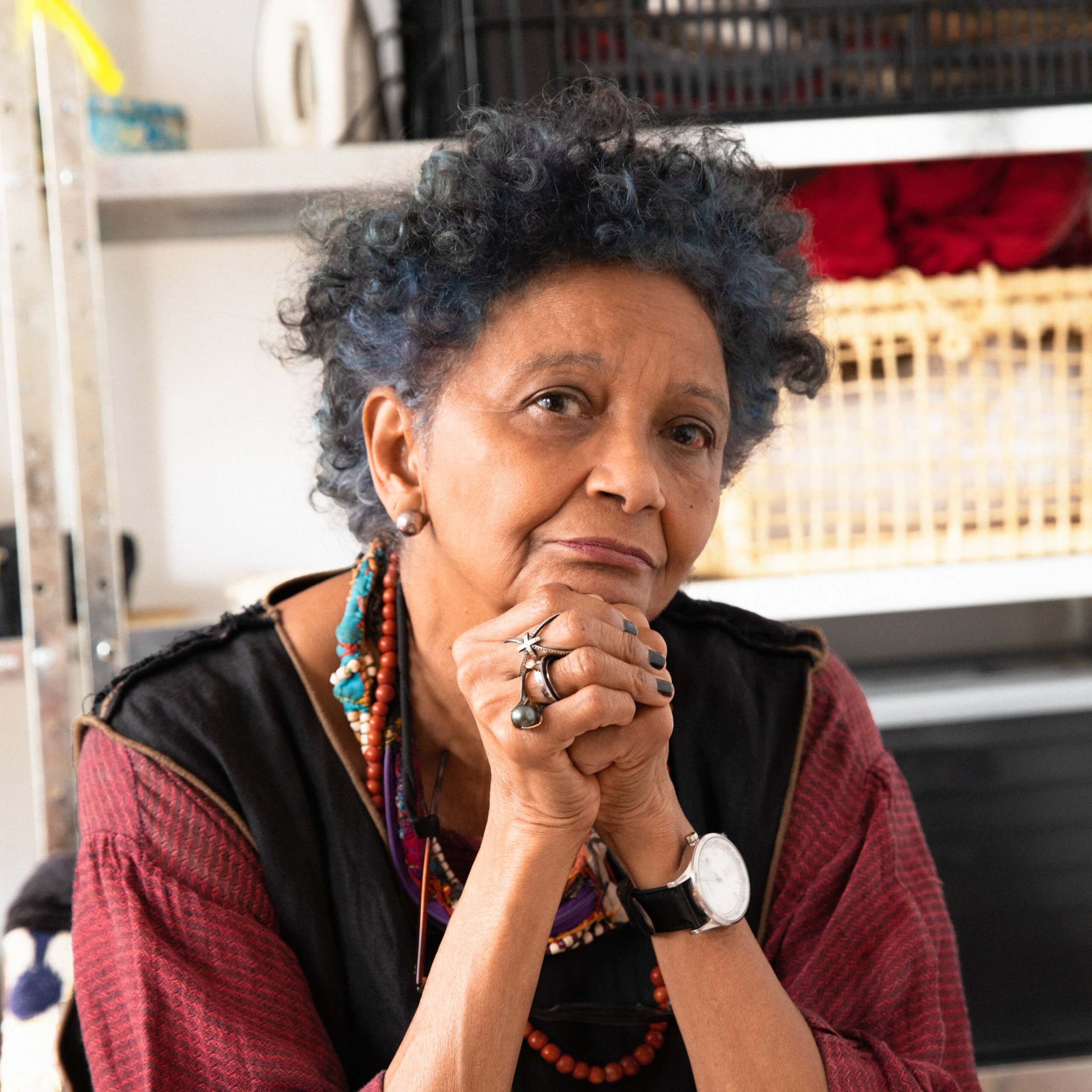 Une femme d'un certain âge, aux cheveux bouclés et parée de bijoux colorés, pose son menton sur ses mains et regarde pensivement l'appareil photo. On aperçoit à l'arrière-plan des étagères sur lesquelles sont disposés divers objets.