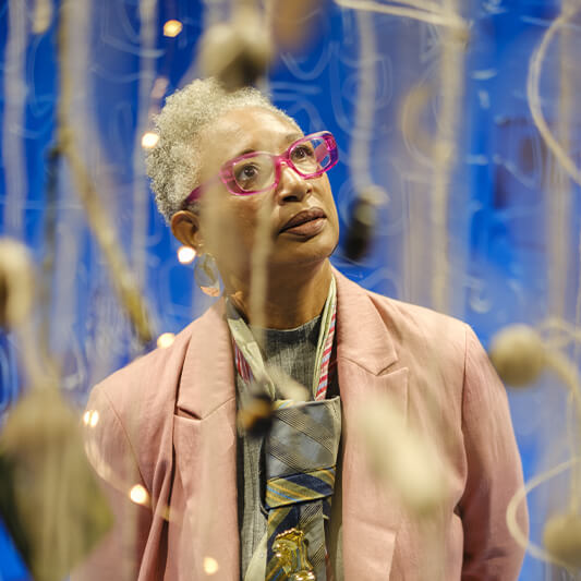 Une personne aux cheveux courts gris et portant des lunettes roses, vêtue d'un blazer rose et d'une écharpe à motifs, observe des objets suspendus sur un fond bleu.
