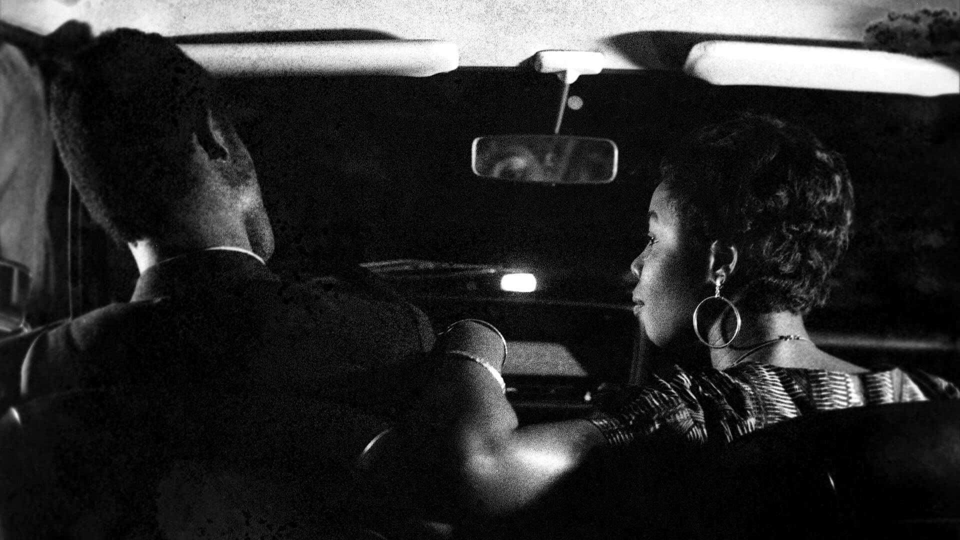 Un homme et une femme sont assis sur les sièges avant d'une voiture, de nuit, vus de dos, la femme regardant vers l'homme.