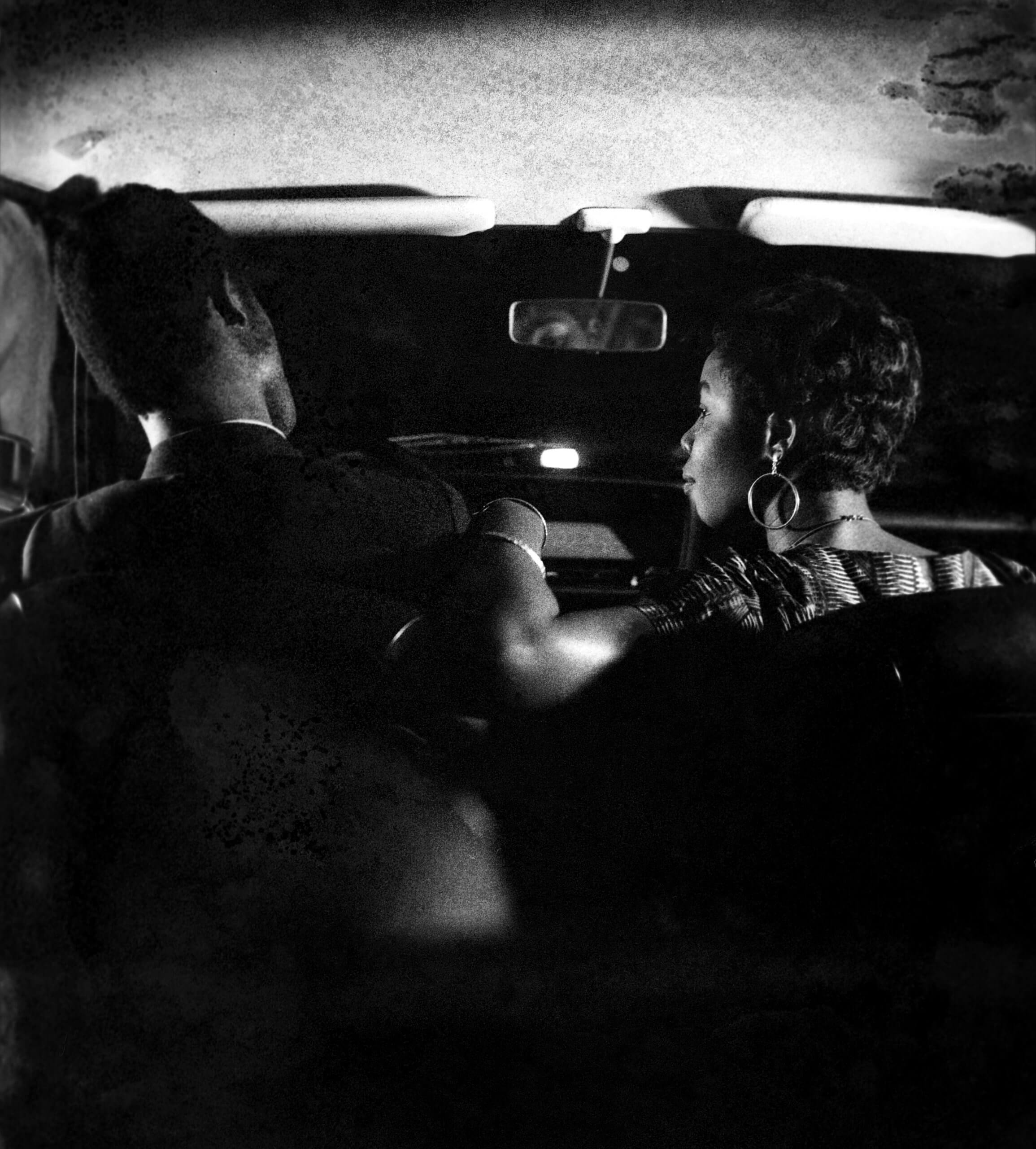 Un homme et une femme sont assis sur le siège avant d'une voiture de nuit, vue depuis le siège arrière, avec le tableau de bord et le rétroviseur visibles.
