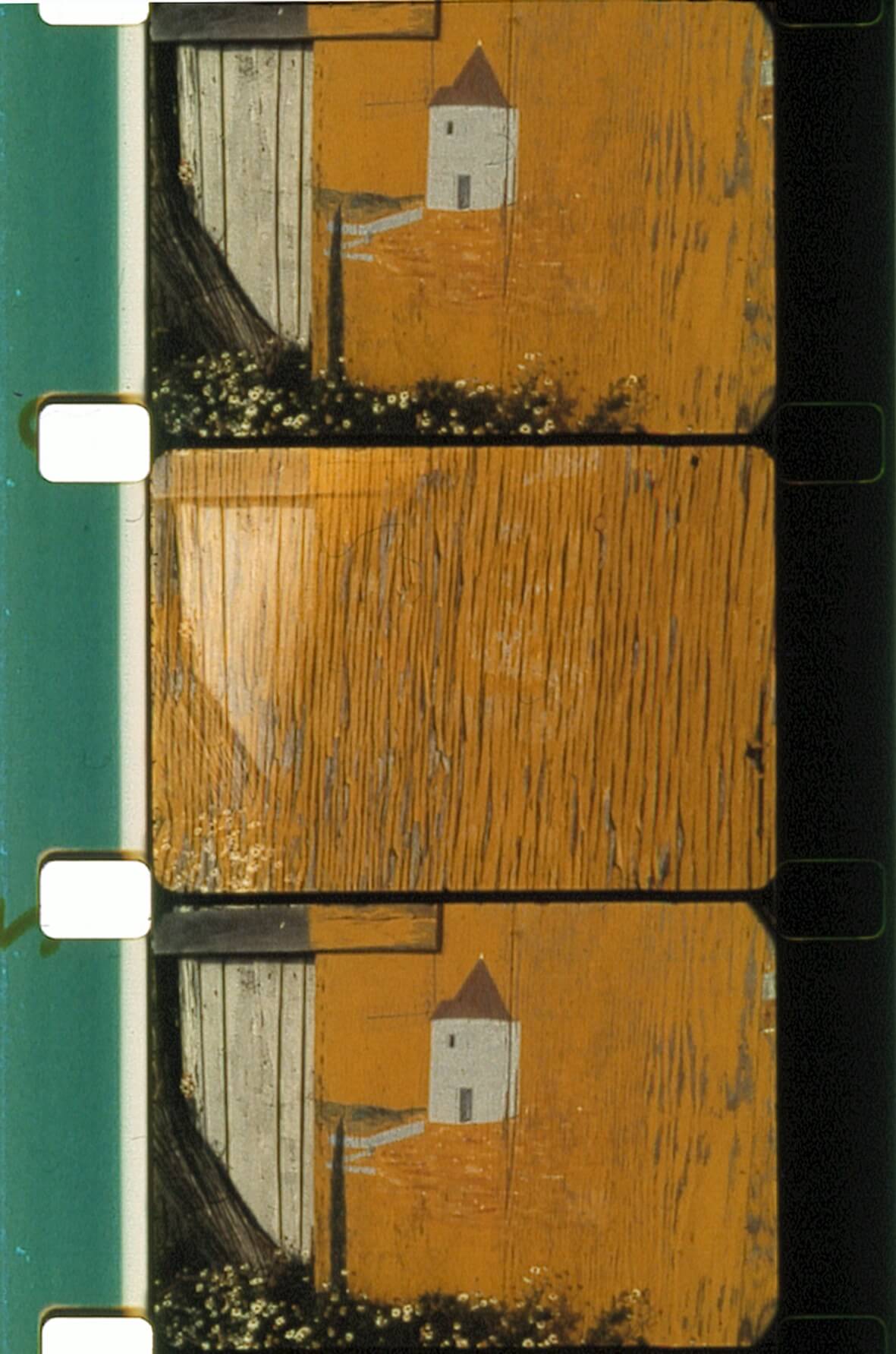 Une bande de pellicule de 35 mm comportant trois images ; l'image du haut représente une scène peinte avec une maison, un arbre et des fleurs, tandis que les deux autres images montrent principalement une texture de bois.