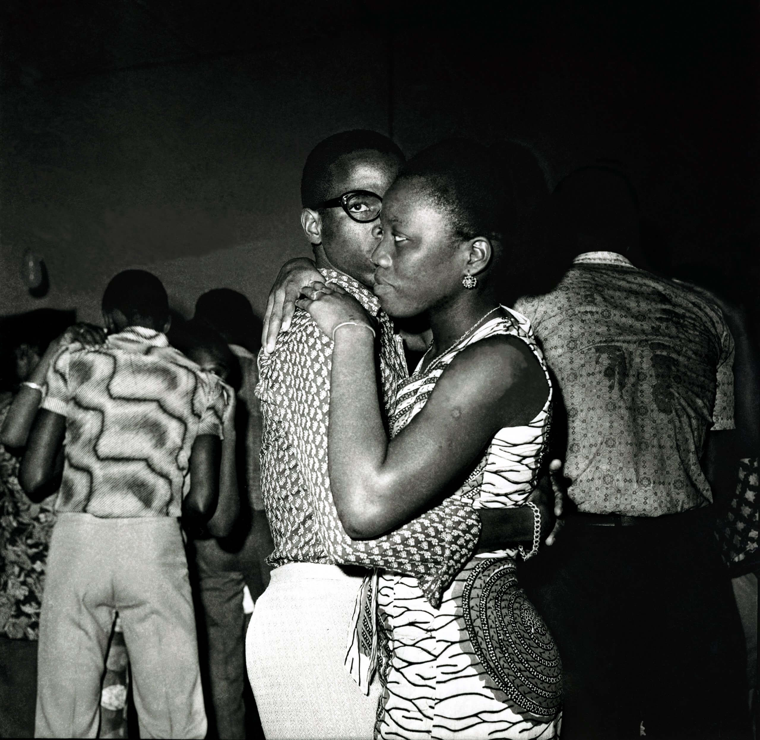 Un couple s'embrasse et danse étroitement lors d'un événement social, entouré d'autres personnes qui dansent à l'arrière-plan.