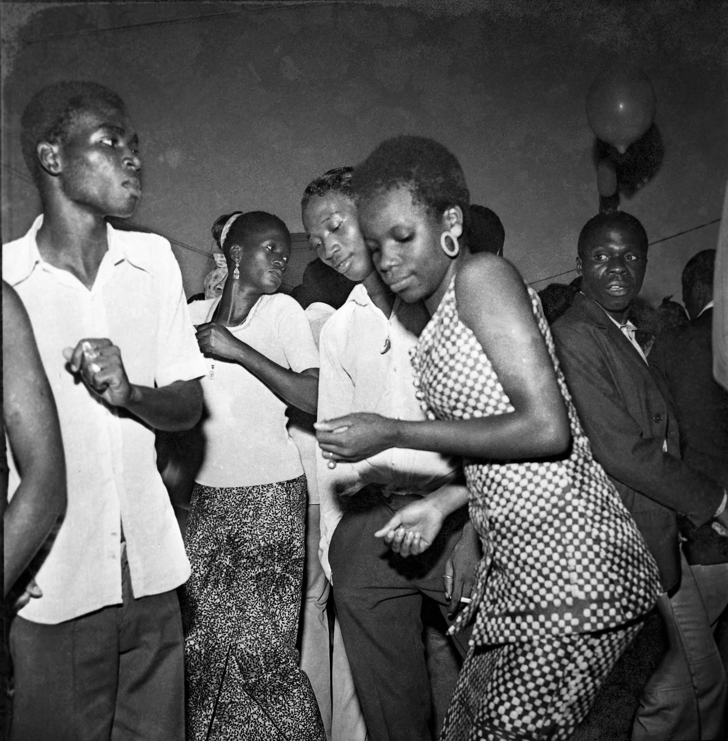 Photo en noir et blanc de jeunes hommes et de jeunes femmes dansant ensemble lors d'une fête ou d'une réunion sociale en intérieur.