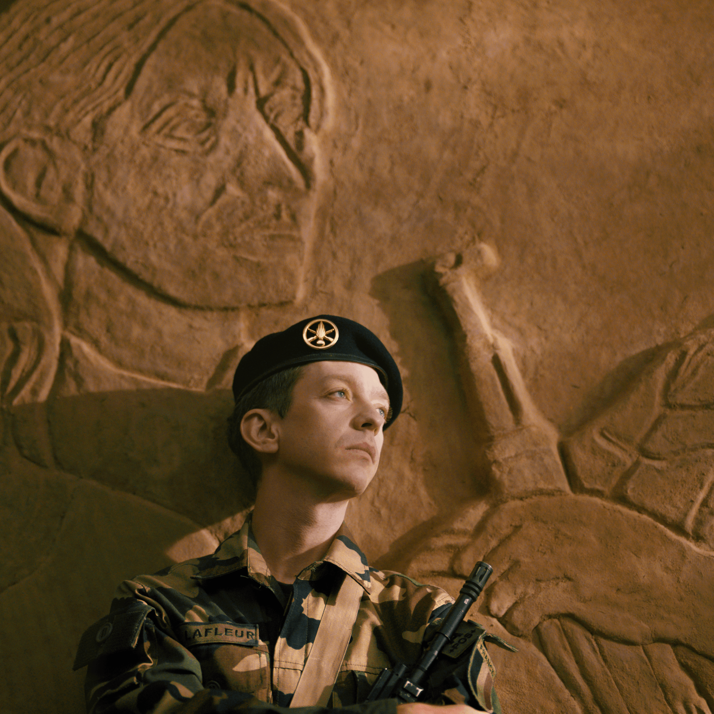 Un soldat en uniforme se tient devant un mur orné d'un grand relief représentant une personne tenant une arme, le regard tourné vers le ciel, l'air pensif.