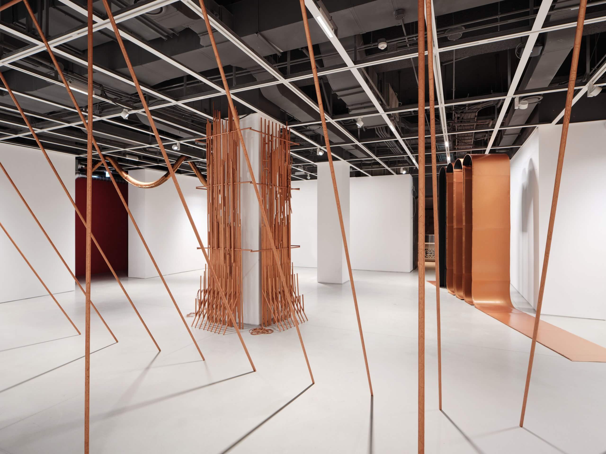 Une galerie d'art contemporain dotée de tiges de cuivre disposées verticalement et en diagonale, d'un pilier central recouvert de lattes de bois, de murs blancs et d'un plafond de style industriel.