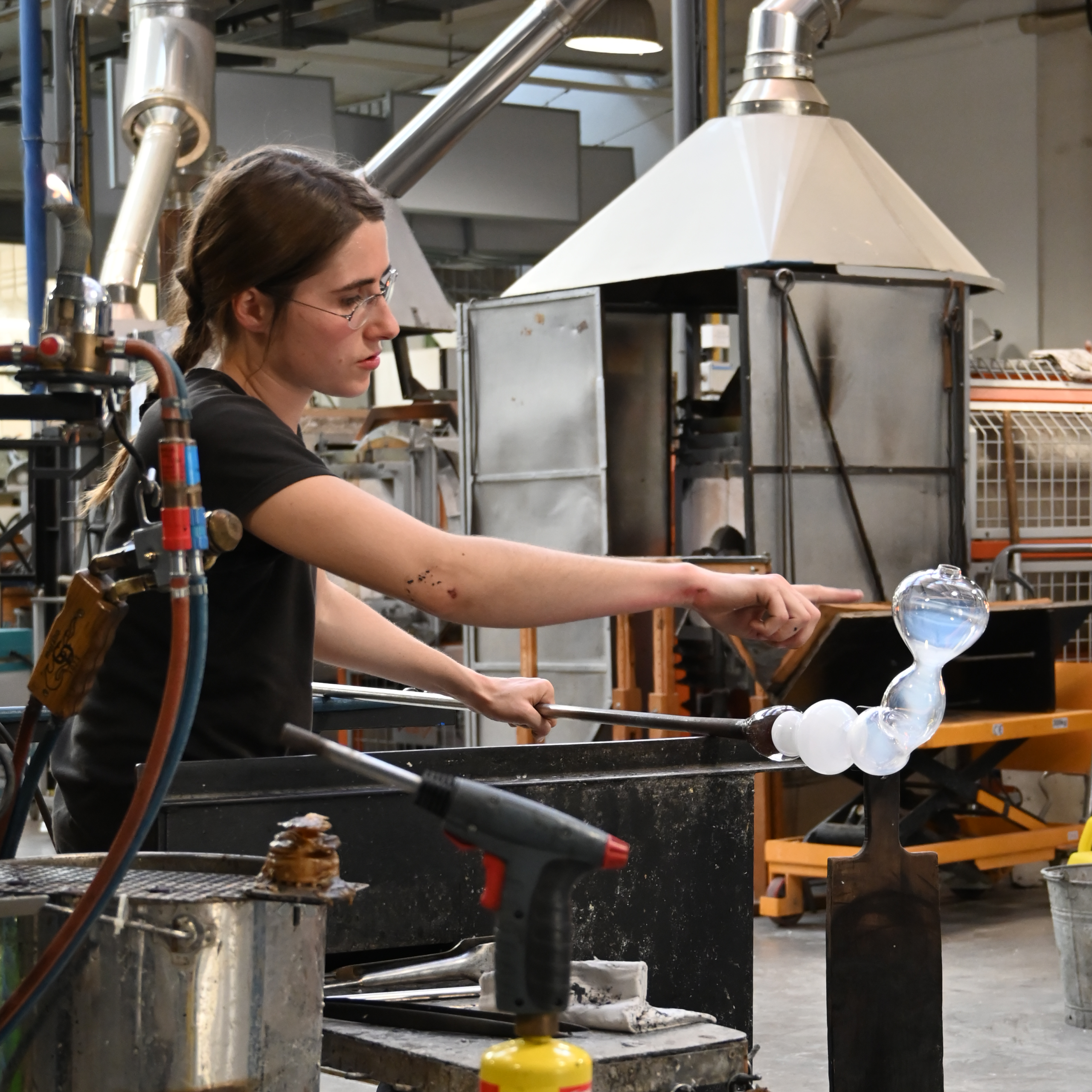 Dans un atelier industriel de soufflage de verre, une personne façonne du verre en fusion à l'aide d'une canne à souffler et d'outils, au milieu d'équipements et de machines.