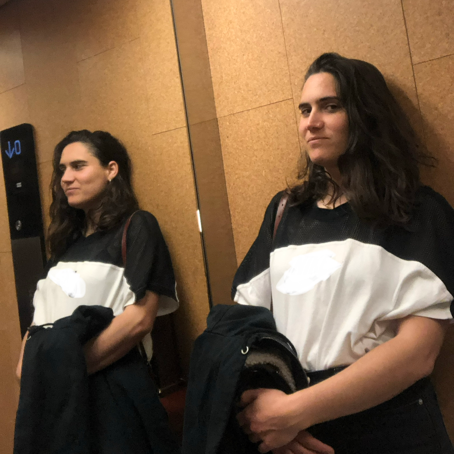 Une femme debout devant un mur en bois et un miroir, tenant une veste, son reflet étant visible à côté d'elle.