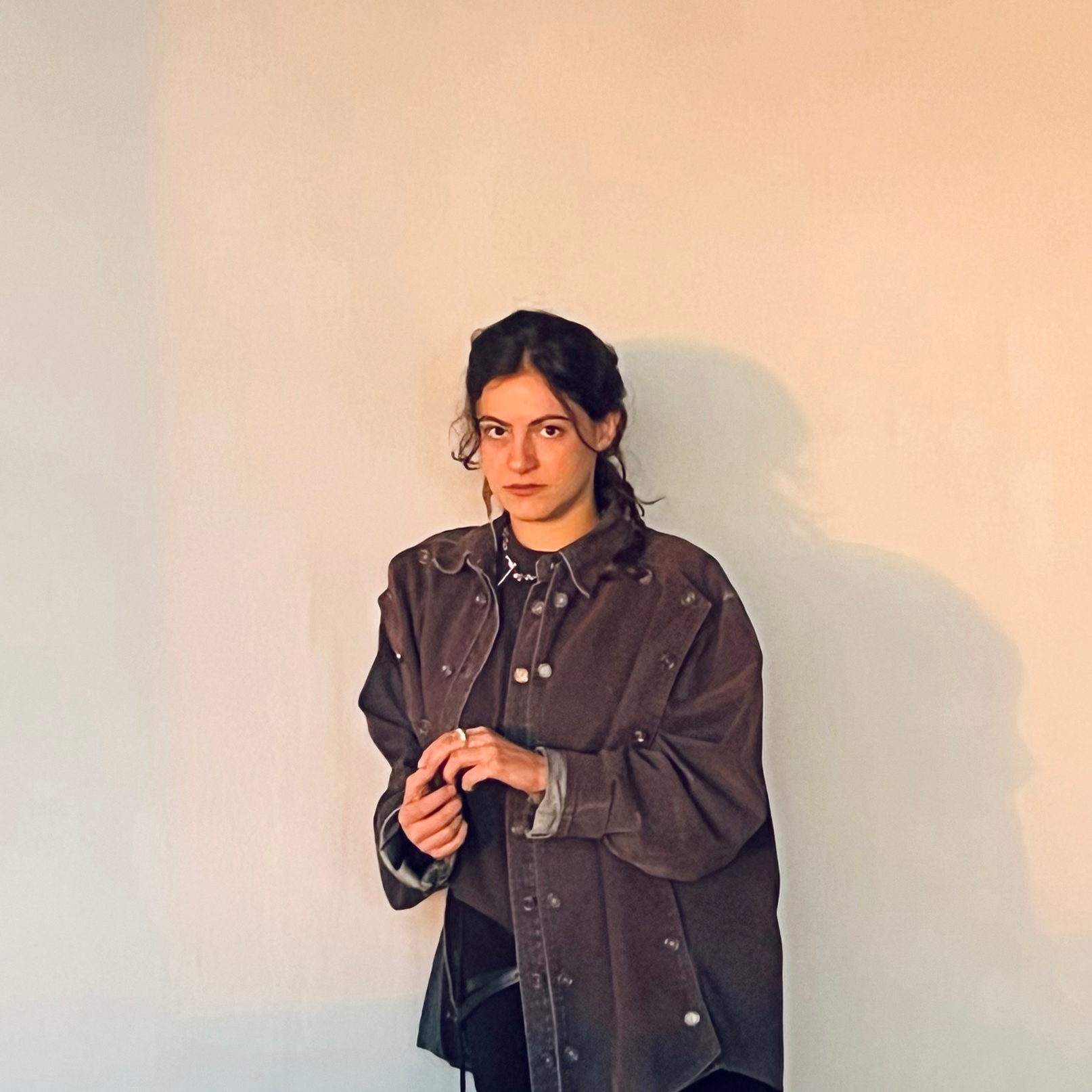 Une femme aux cheveux foncés attachés en arrière se tient debout devant un mur uni de couleur claire ; elle porte une veste foncée boutonnée par-dessus une chemise foncée et regarde l'appareil photo avec une expression neutre.